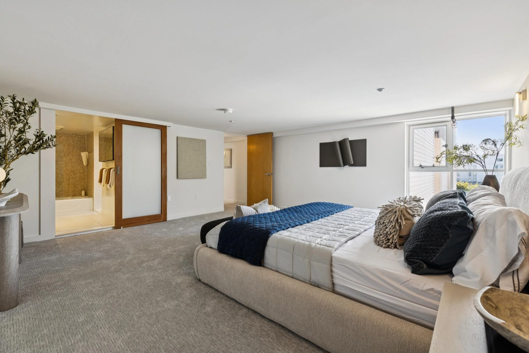 A bedroom with a bed covered in a beige and dark blue quilt, numerous pillows, a wall-mounted flat-screen TV, a large window with a city view, a potted plant, and an open door leading to a bathroom.