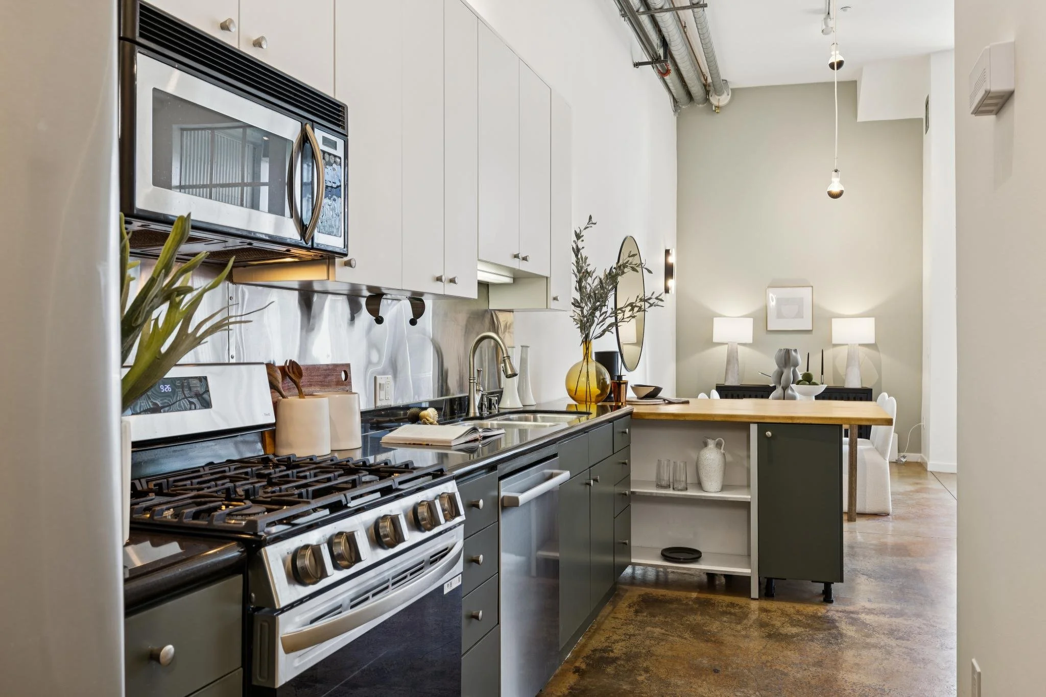 Modern kitchen with white cabinets, stainless steel appliances, a black stove, a wooden countertop island, decorative vases, and a dining area with lamps and artwork in the background.