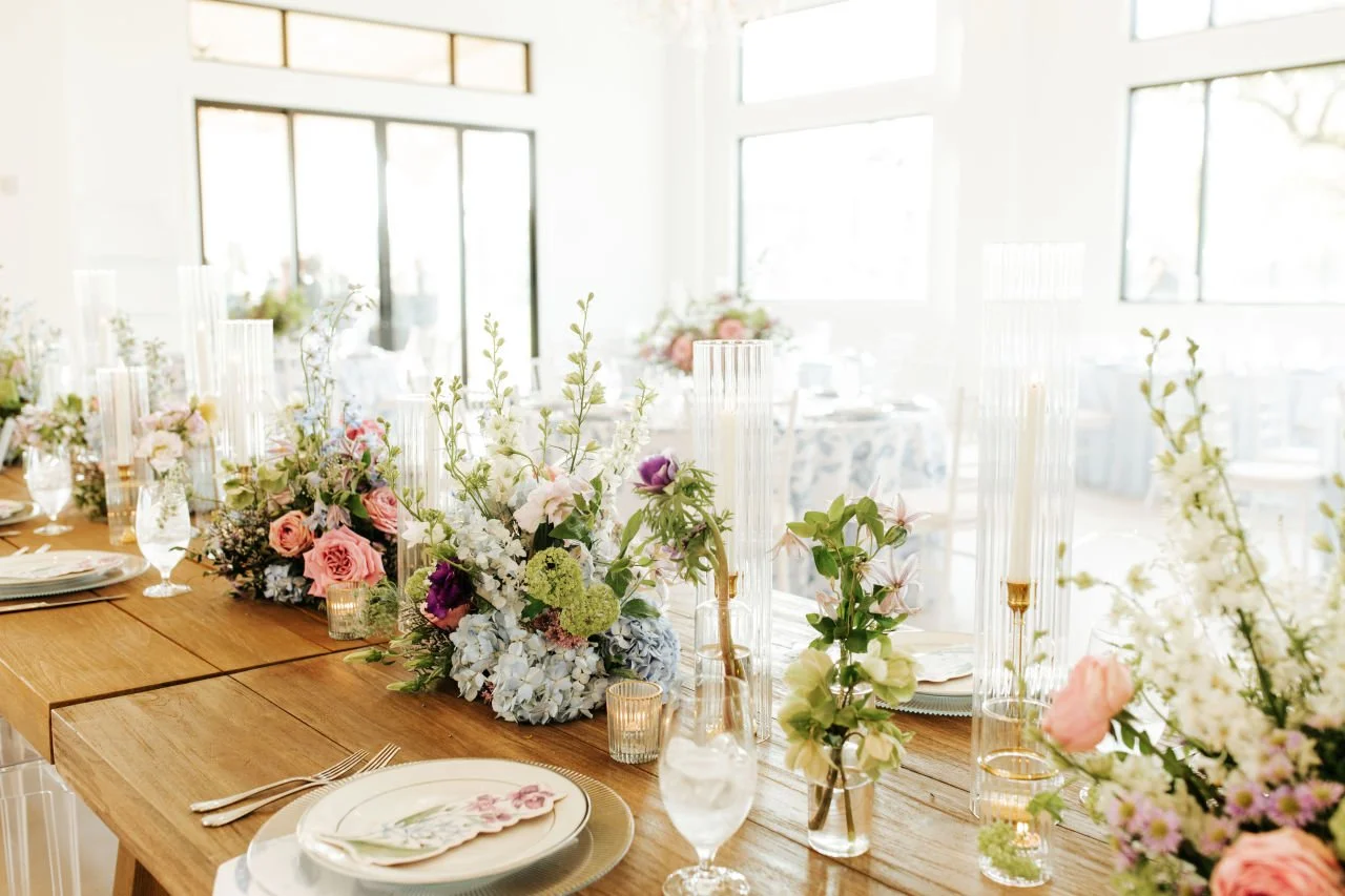 Elegant wedding reception table with floral centerpieces, custom menu cards, tall candles, and place settings in a bright, airy room with large windows.