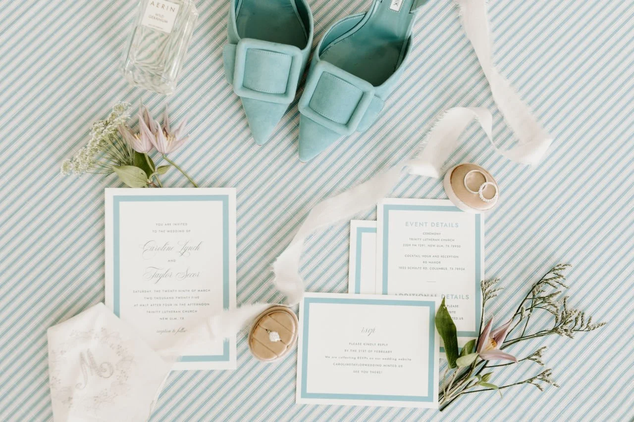 Wedding invitation cards, mint green shoes with bows, a bottle of perfume, flowers, rings, and decorative fabrics on a striped tablecloth.