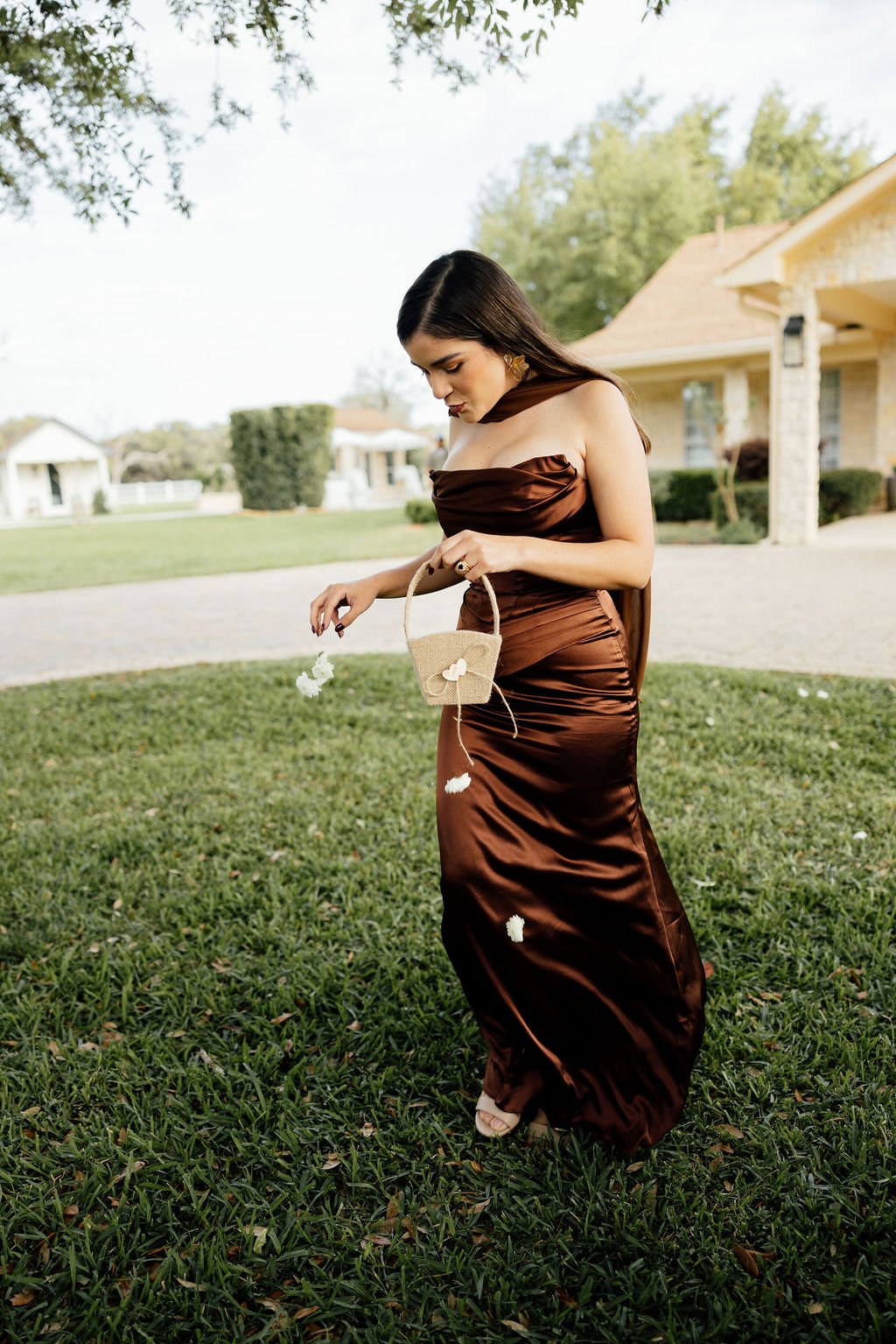 A woman in a brown satin dress standing outdoors on green grass, holding a small beige handbag with white flower decorations, surrounded by trees. Outside in the day time at a wedding ceremony.