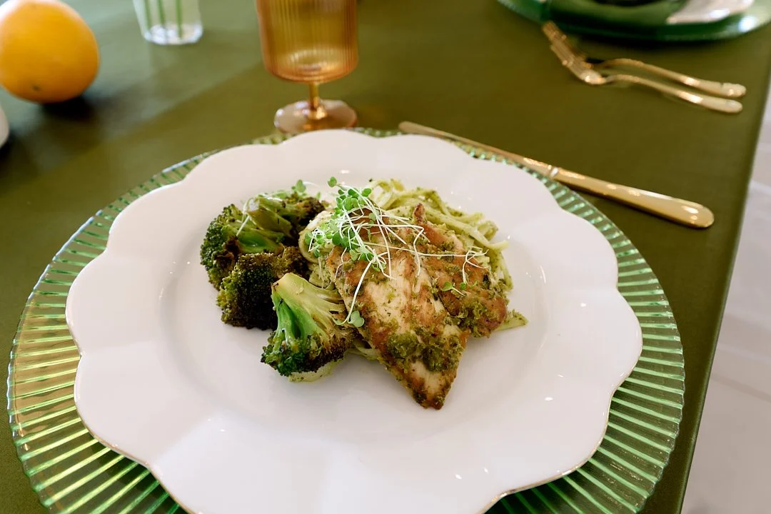 A plate of baked fish topped with herbs and microgreens, served with broccoli, on a decorative white and green plate at a dinner table.