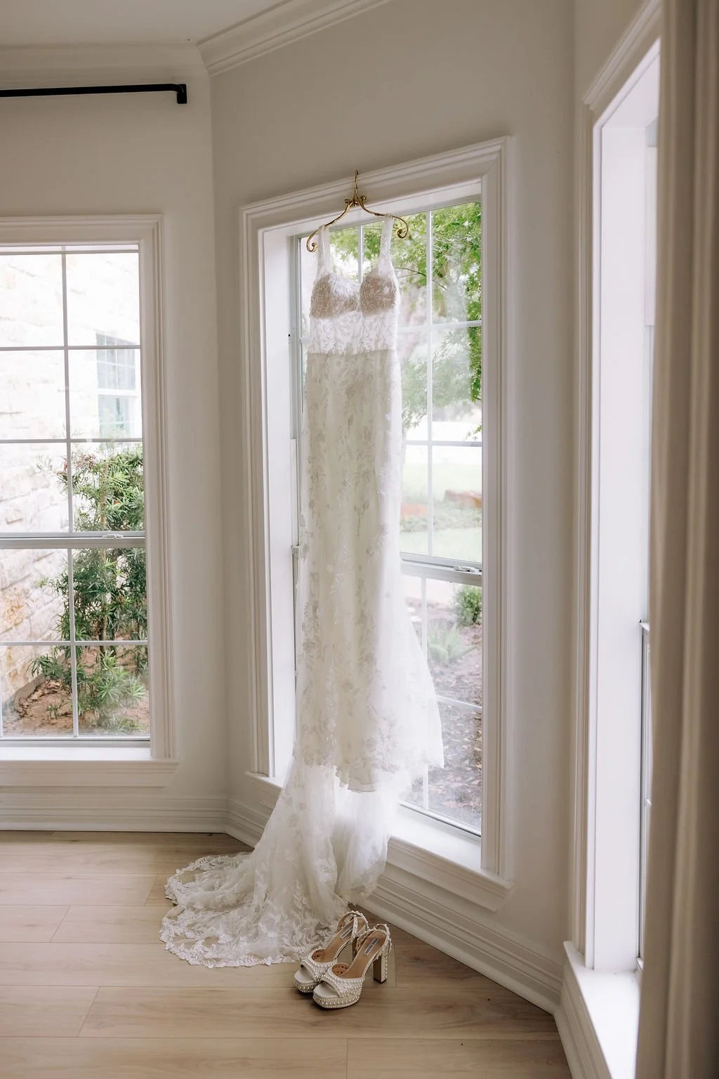 A white lace wedding dress hanging on a gold hook in front of a large bay window, with a pair of white high-heeled shoes with rhinestones placed on the floor beneath the dress.