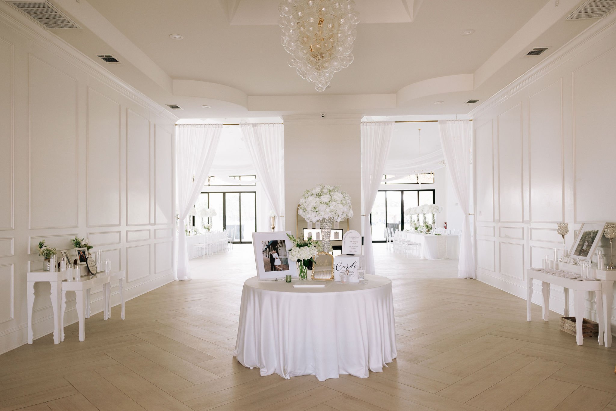 Elegant white decorated room with a round table in the center, floral arrangements, framed photos, and a chandelier