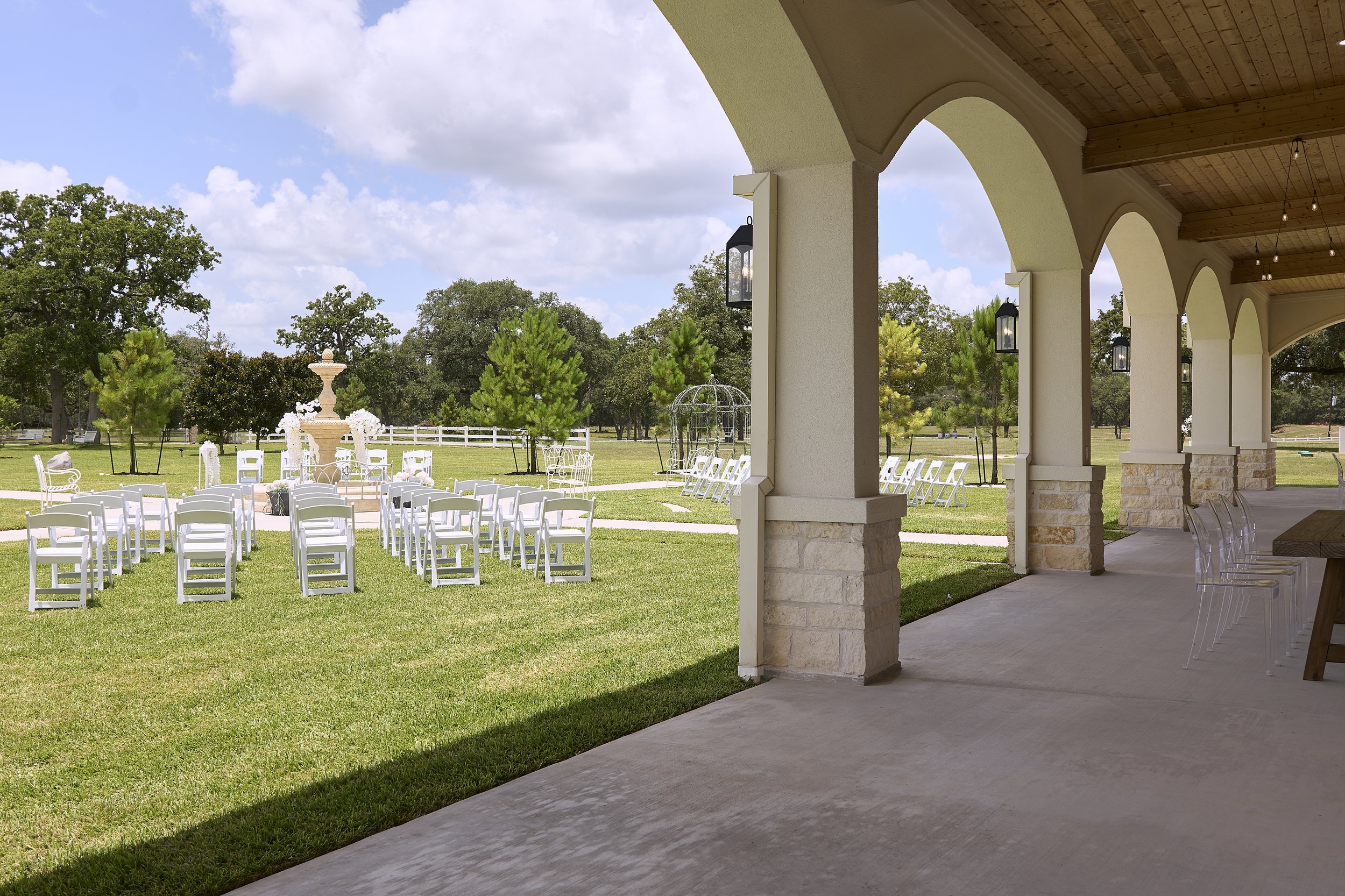 Outdoor wedding setup with white chairs arranged in rows on a grassy lawn, a fountain, trees, and a partly cloudy sky, viewed from a covered patio with hanging light bulbs.