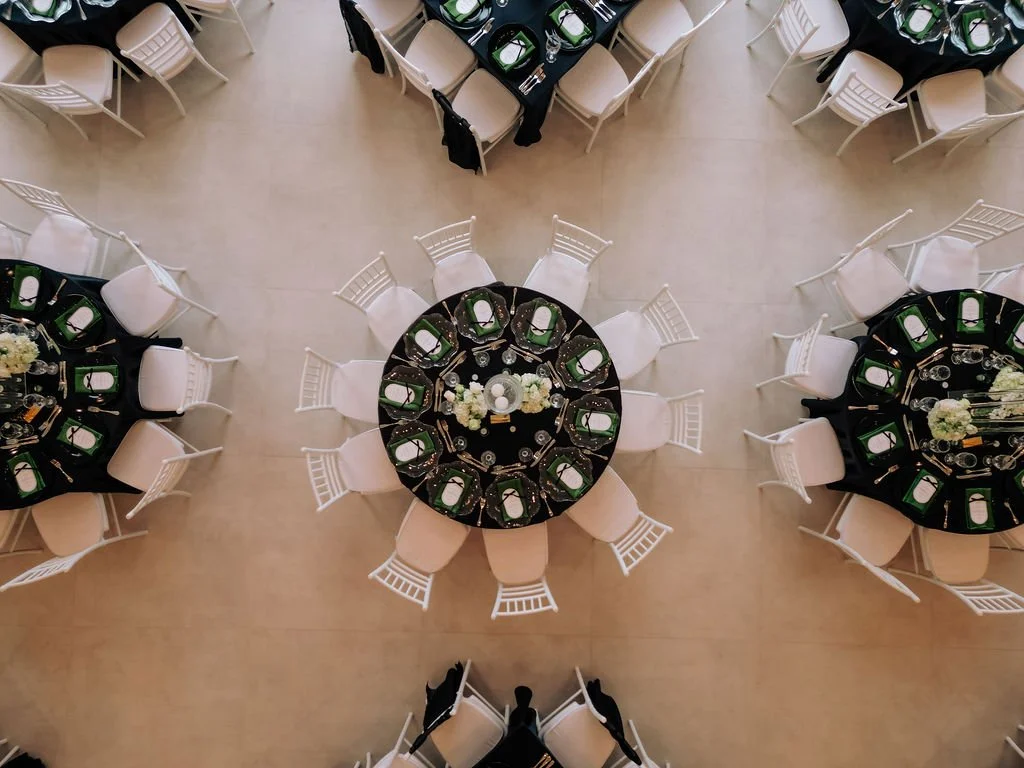 Overhead view of a wedding receptionl with three round tables set. Each table is covered with a black tablecloth, decorated with white floral centerpieces, and set with plates, glasses, silverware, and place cards. White chairs surround each table.