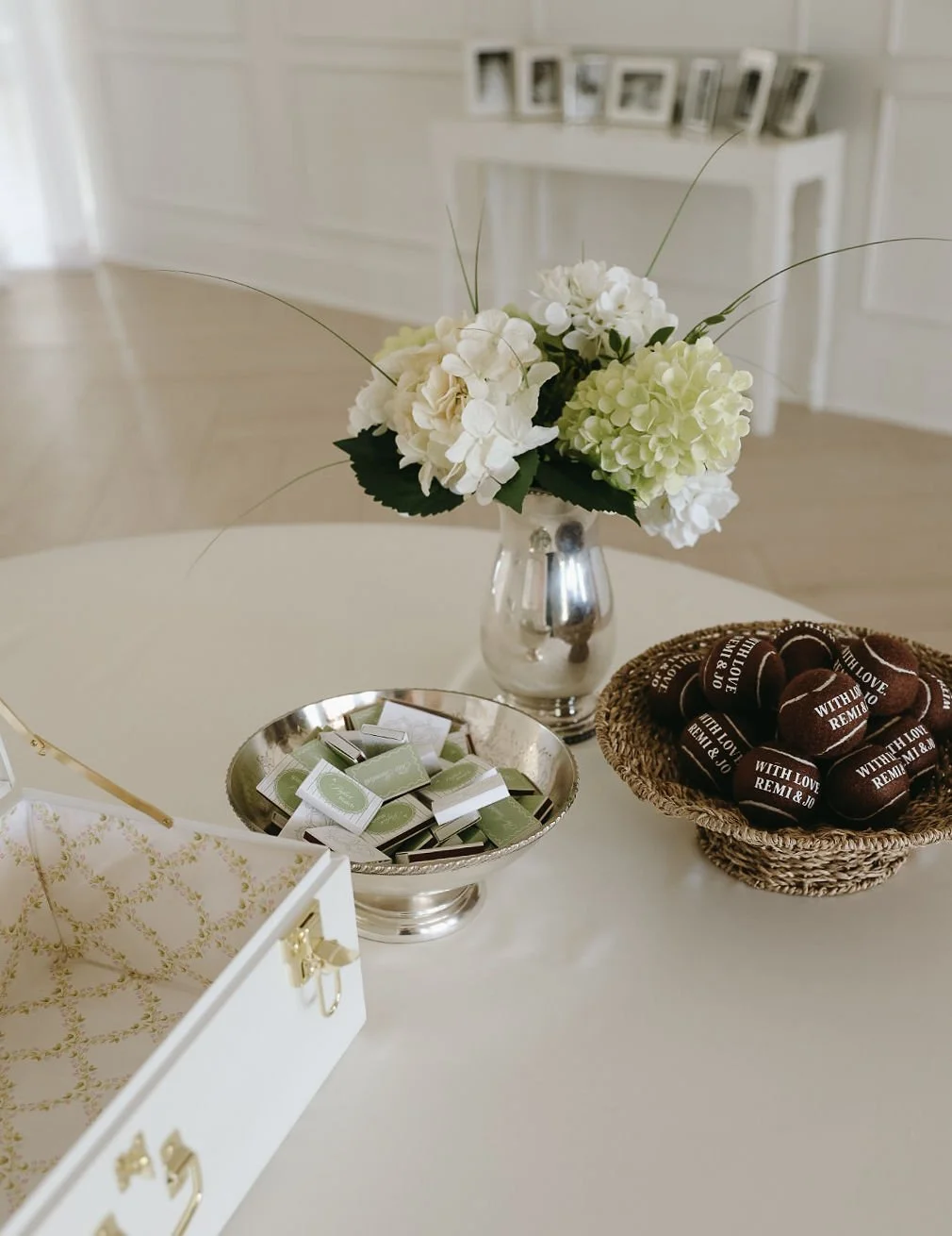 A round table with a white tablecloth, decorated with a flower arrangement of white and pale green hydrangeas in a silver vase, a silver bowl filled with small green and white wrapped items, a woven basket containing chocolate candies wrapped in brow