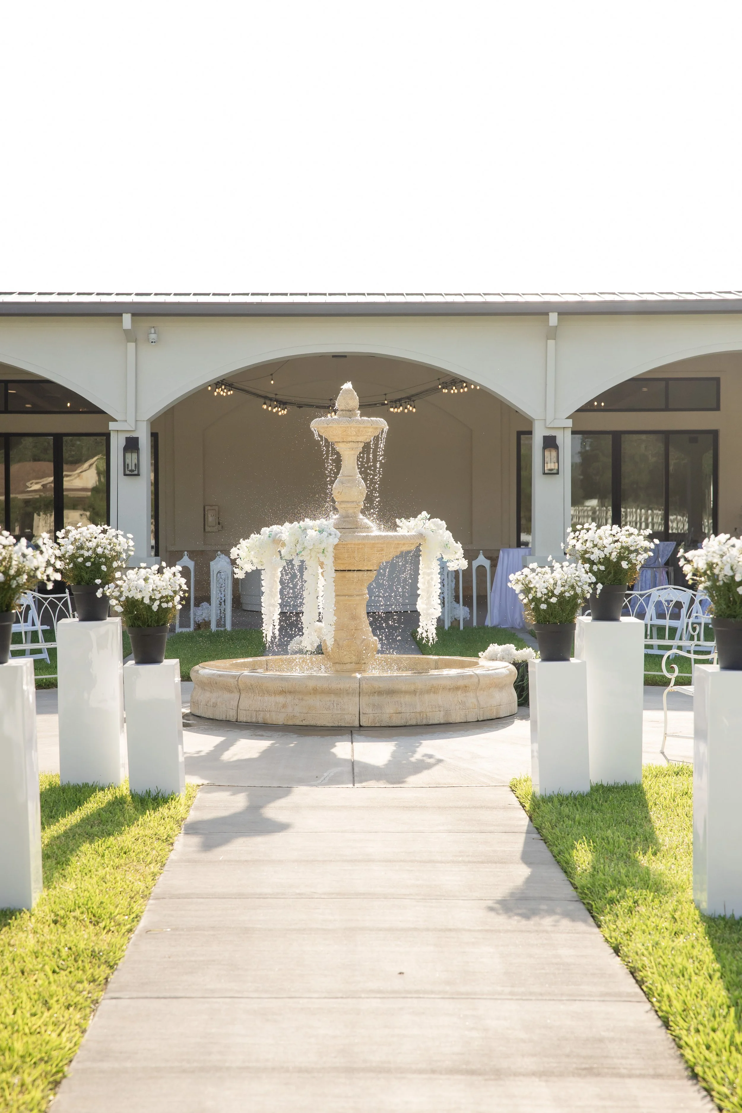 A pathway leading to a white building with arched entryways, featuring a fountain with water flowing and surrounded by white flowers and potted plants.