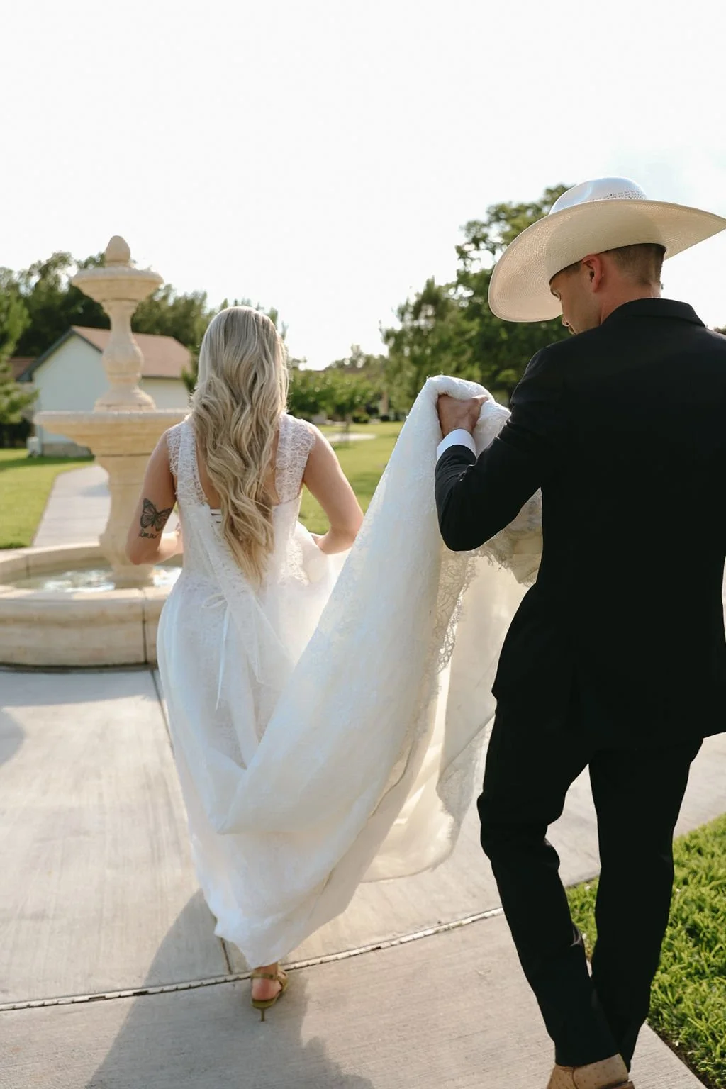 A bride in a white wedding dress with long blonde hair walking away from a groom wearing a black suit and white cowboy hat, holding her wedding dress train, near a fountain and greenery at Ro Manor.