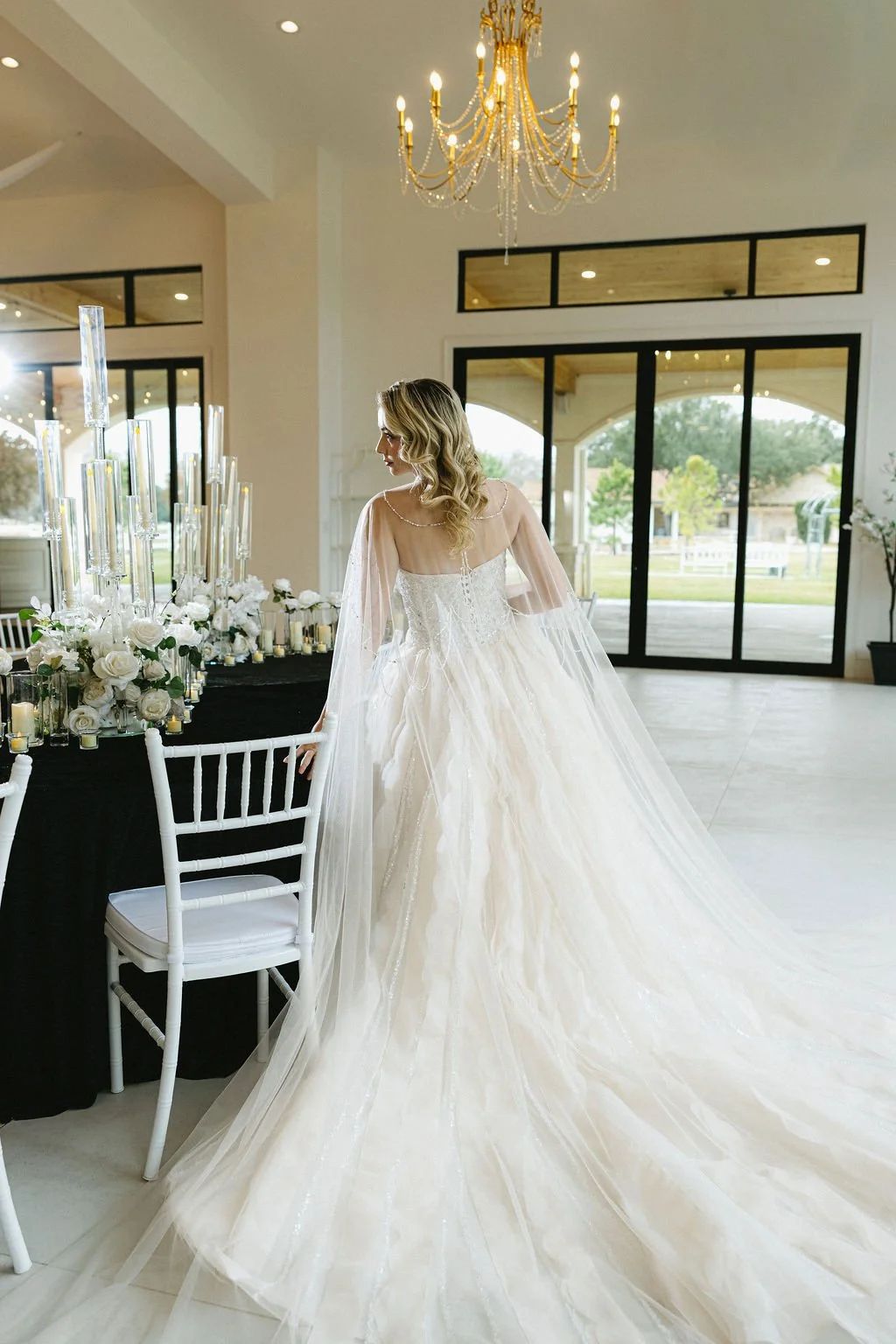 A bride in a white wedding dress with a long train stands in a decorated event hall with large windows and a chandelier, looking at a table with white flowers and tall candles.