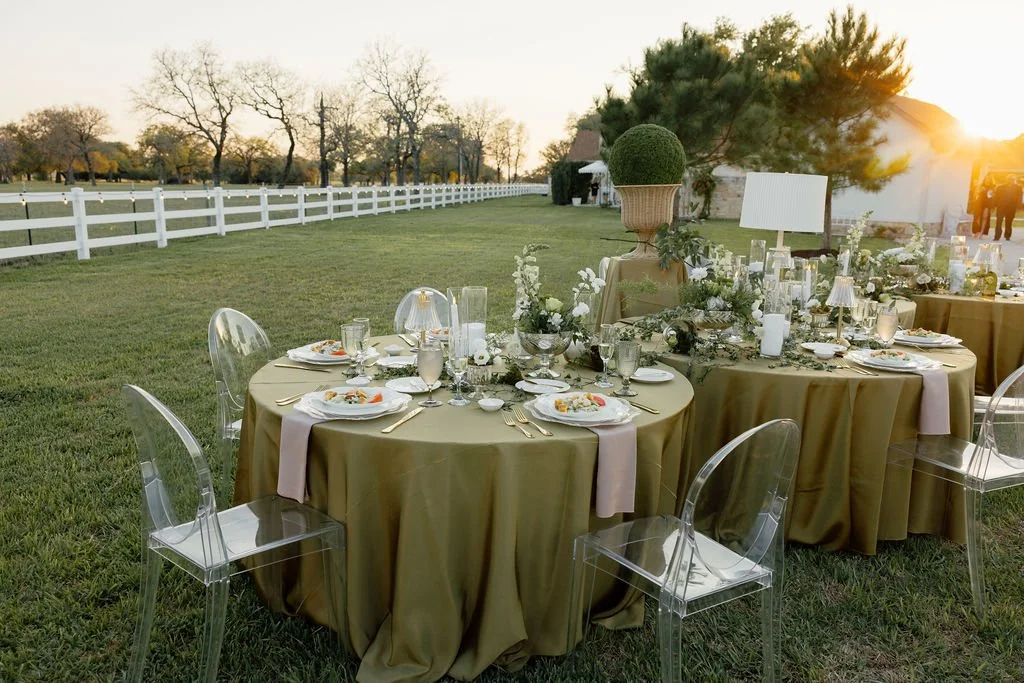 Outdoor wedding reception table set with gold tablecloths, glassware, plates, and silverware, decorated with white flowers and greenery, with clear acrylic chairs and a white fence on a grassy field at sunset.
