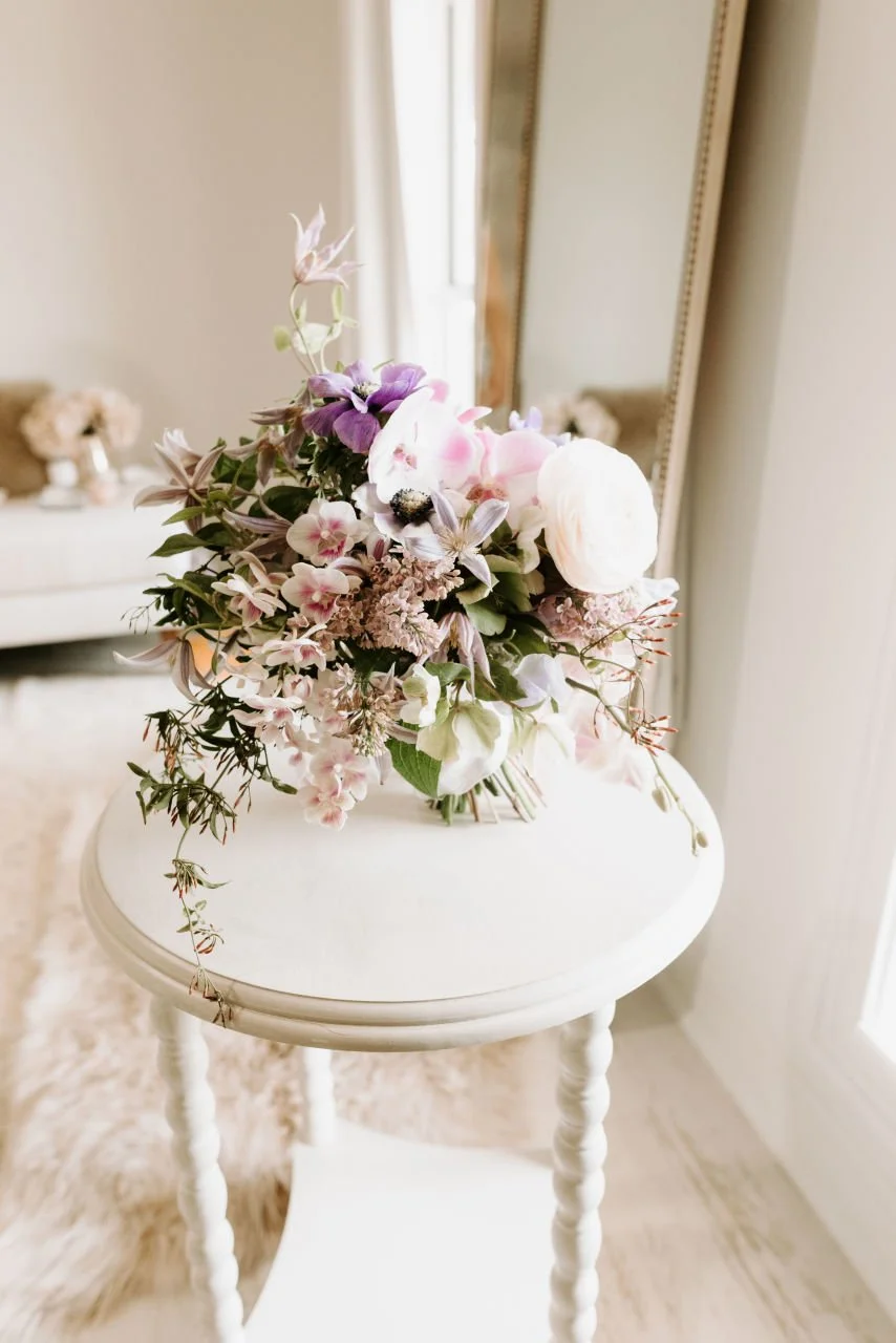 A bouquet of pastel-colored flowers on a white wooden side table in a bright, softly lit room.