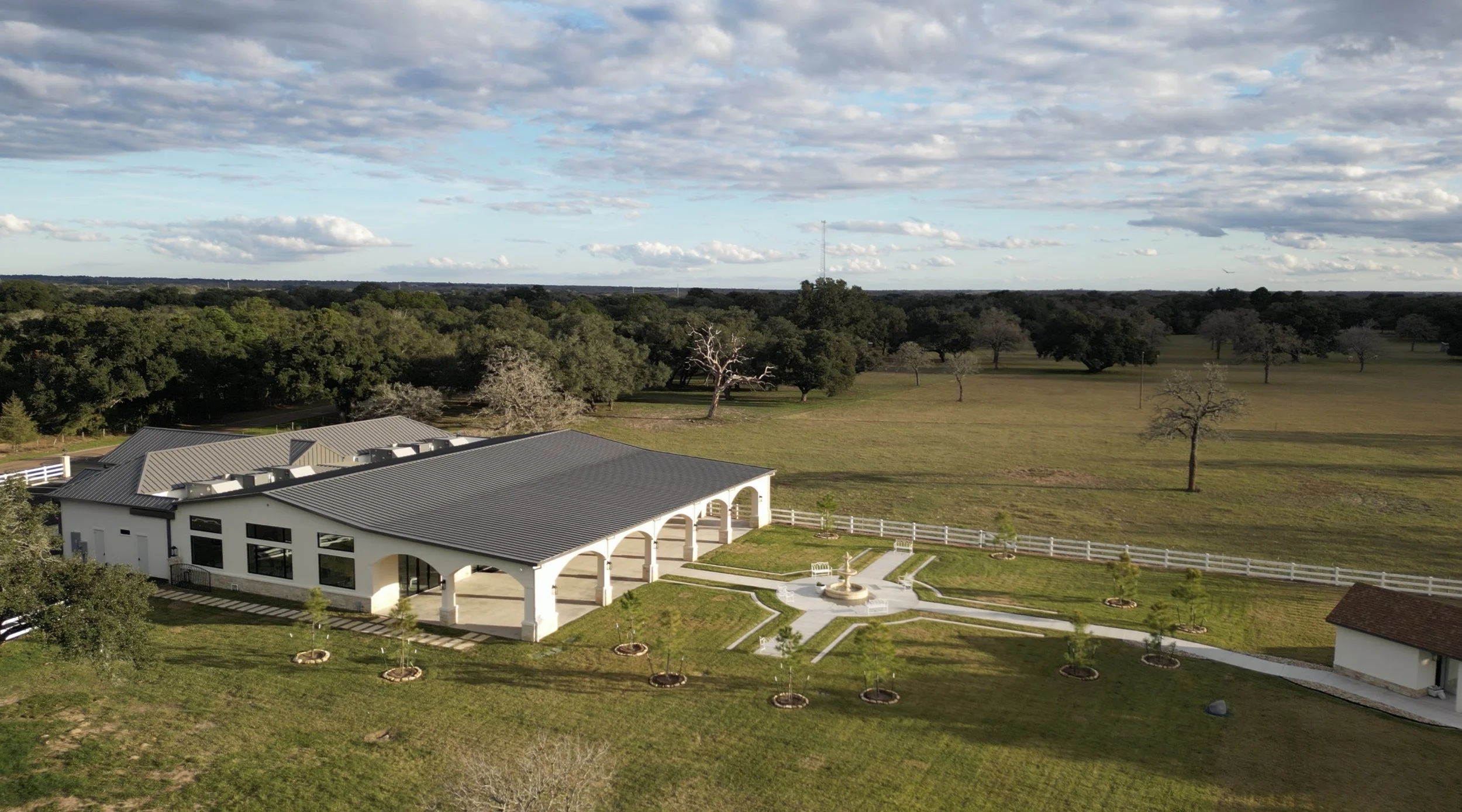 A white house with arches and a fountain in the front yard, overlooking a wide grassy field with scattered trees and a forested background under a partly cloudy sky.