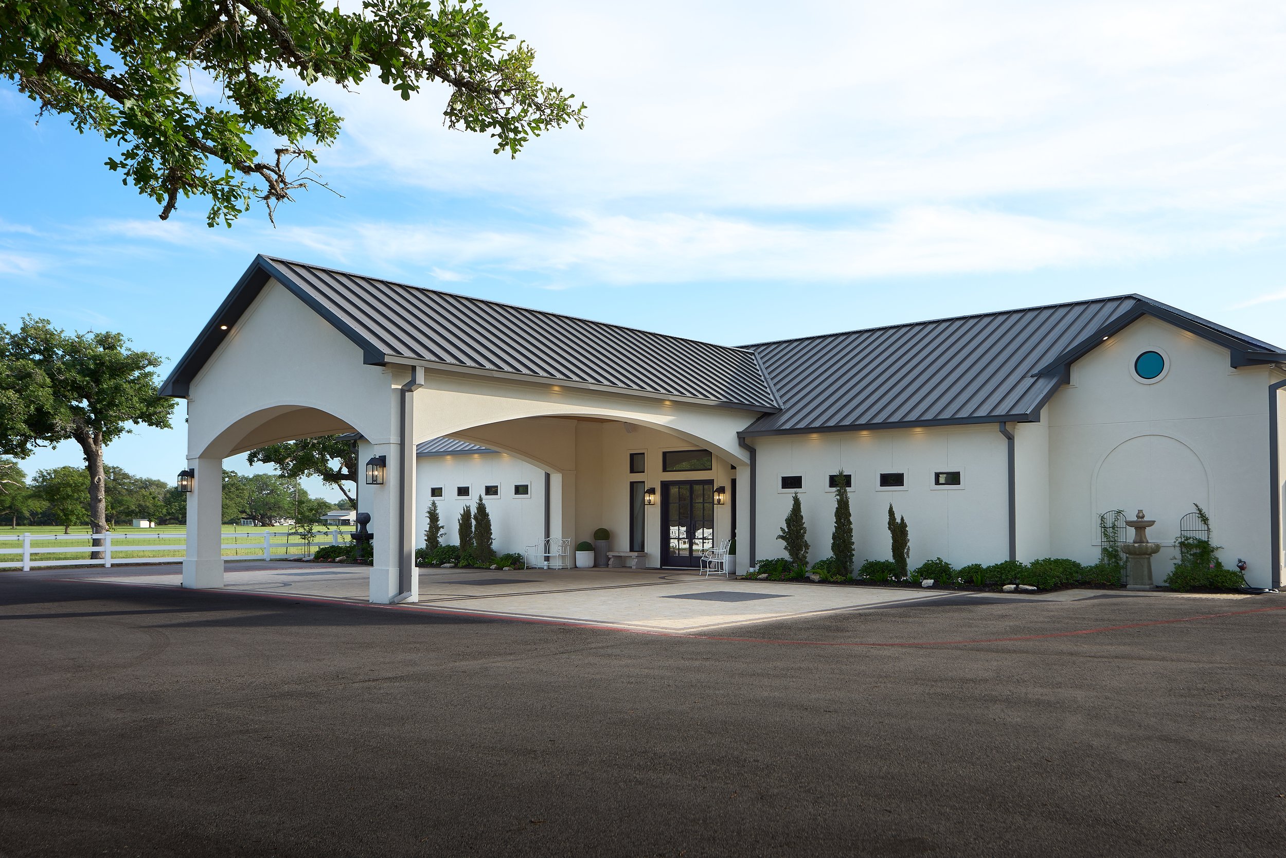 A modern white building with a dark gray metal roof and arched entrance, surrounded by small trees and shrubs, with a large parking lot in the foreground and a partly cloudy sky overhead.