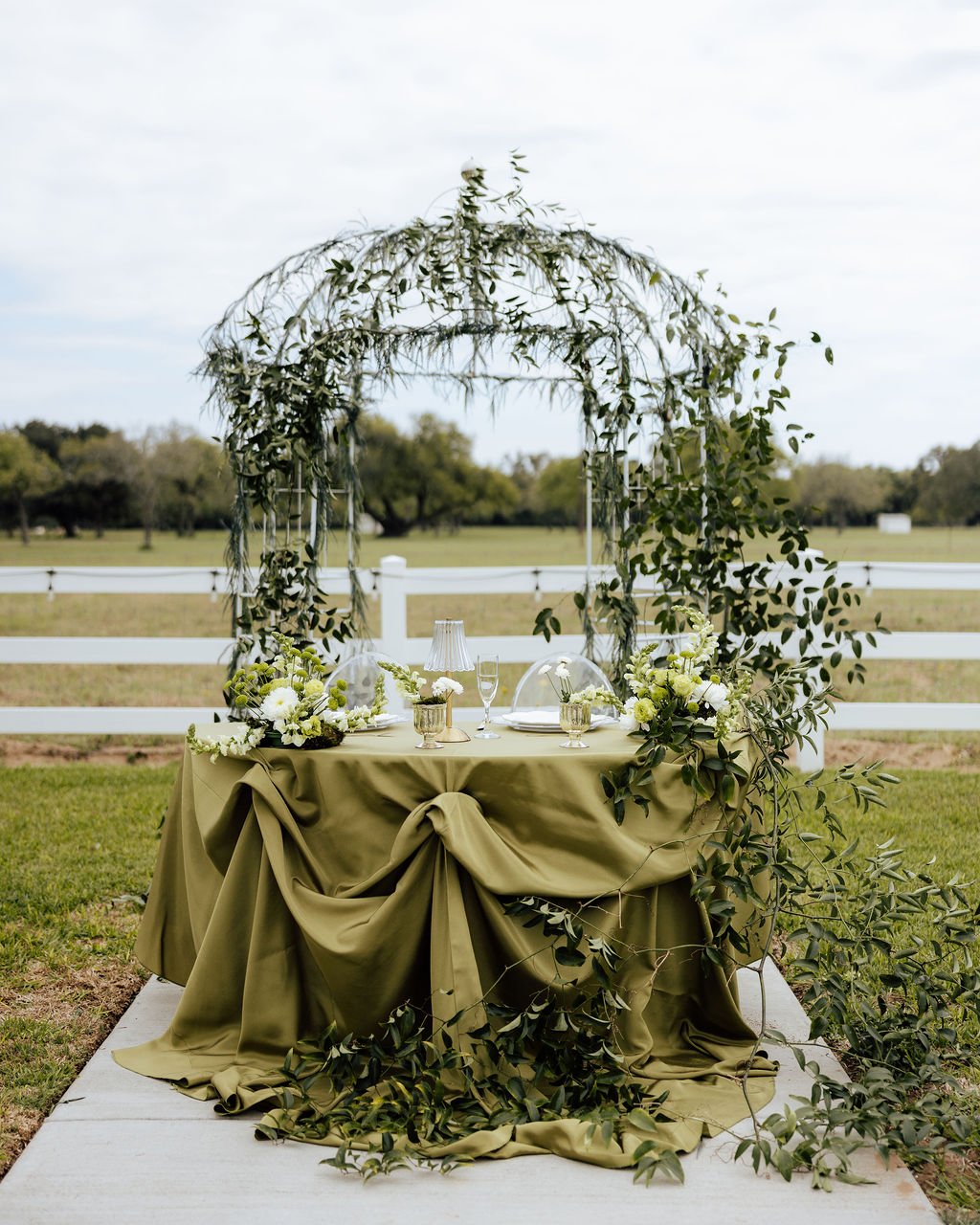 Wedding sweetheart table with green tablecloth under a floral arch outdoors.