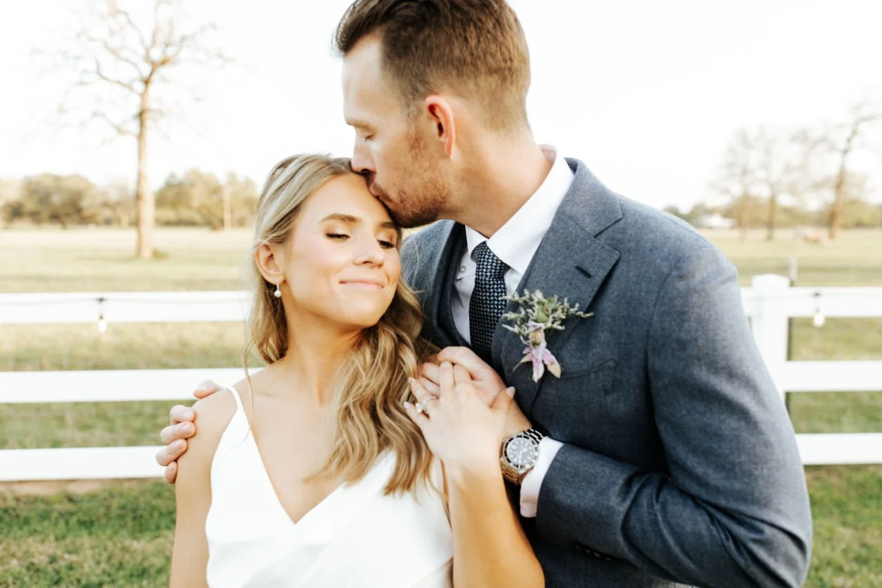 The groom in a suit kisses his newlywed on the forehead outdoors during sunset. The bride wears a white dress and has long wavy hair, standing in front of a white fence with trees in the background.