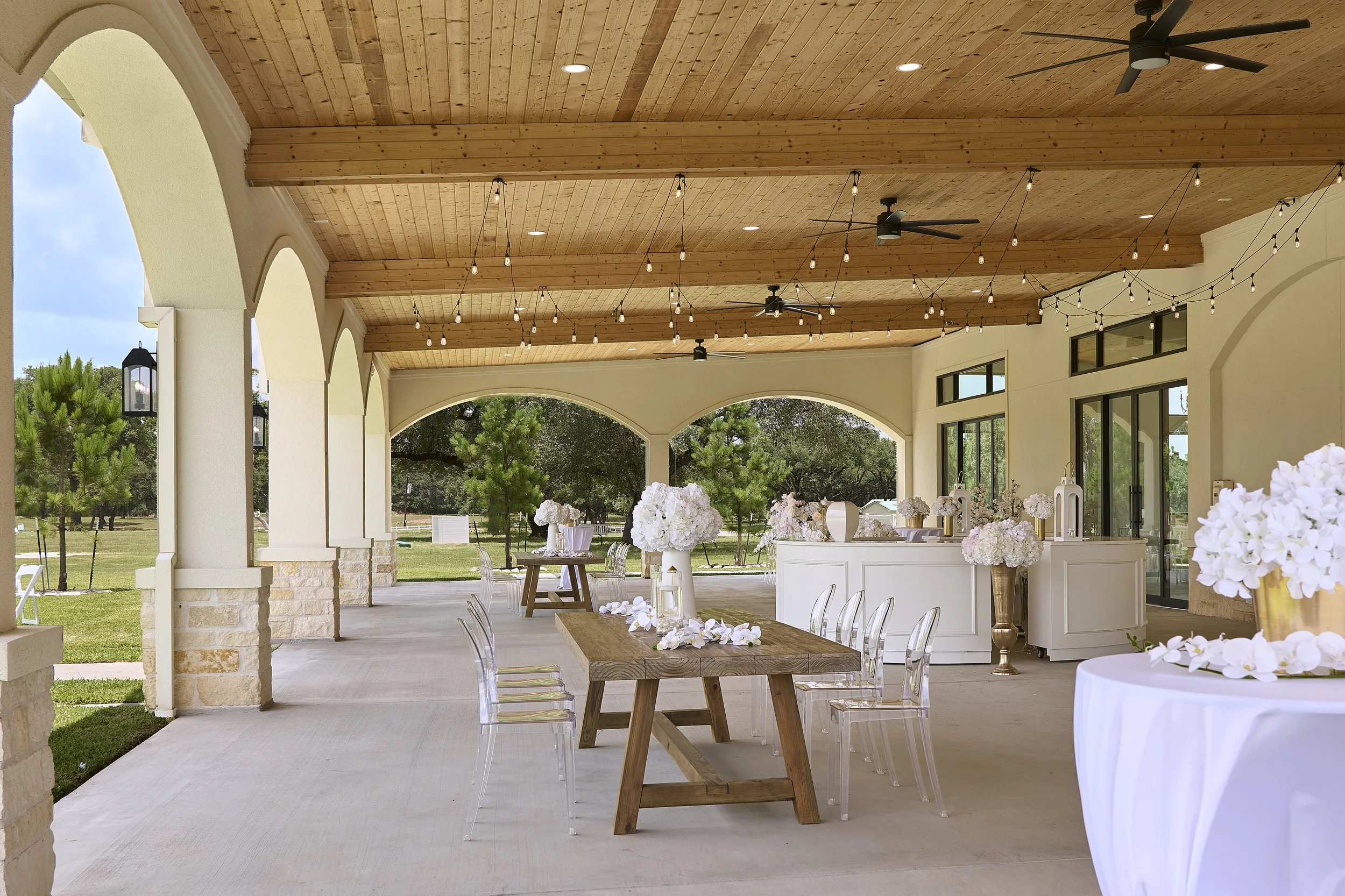 Outdoor event space with wooden ceiling, string lights, ceiling fans, large windows, and tables decorated with white flowers