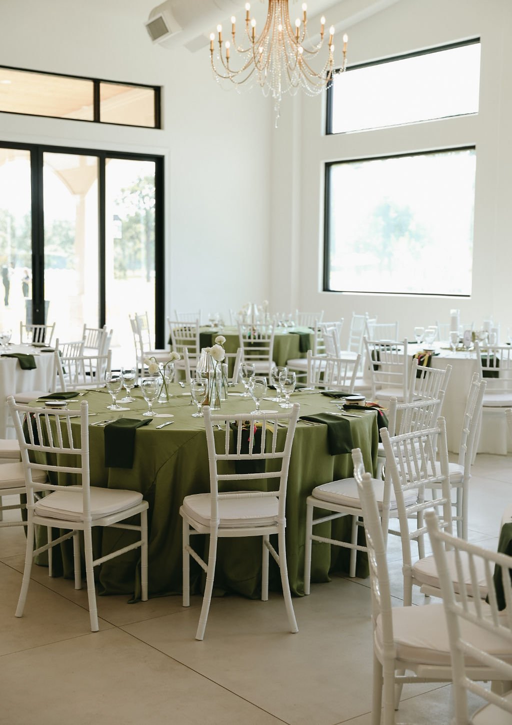 Reception hall with large windows, round tables covered in green tablecloths, white chairs, glassware, and a gold chandelier with crystals hanging from the ceiling.