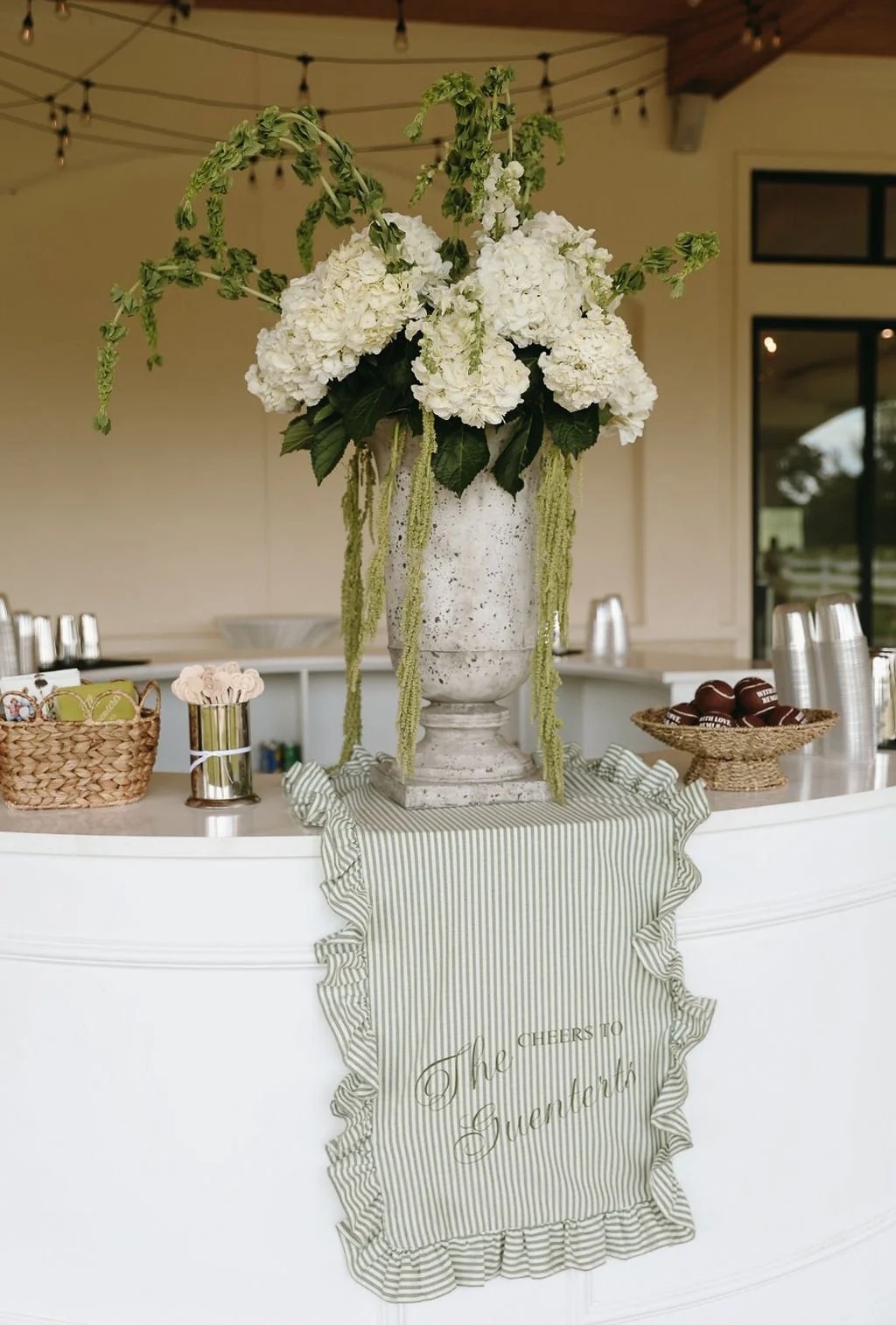 Wedding bar. Large white flower arrangement with hydrangeas in a tall white vase on a curved white counter, with a striped green and white cloth and a sign that reads 'Cheers to The Guenterts'. 