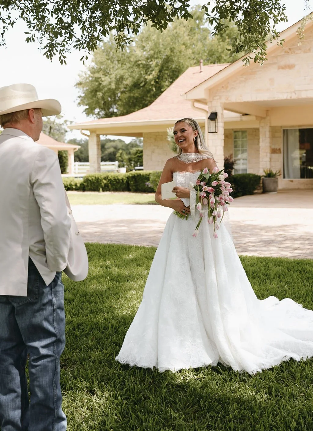 Bride's first look with dad. A bride in a white wedding gown holding a bouquet of pink tulips, smiling and talking to her dad wearing a white blazer, jeans, and a cowboy hat on a lawn outside a house. 