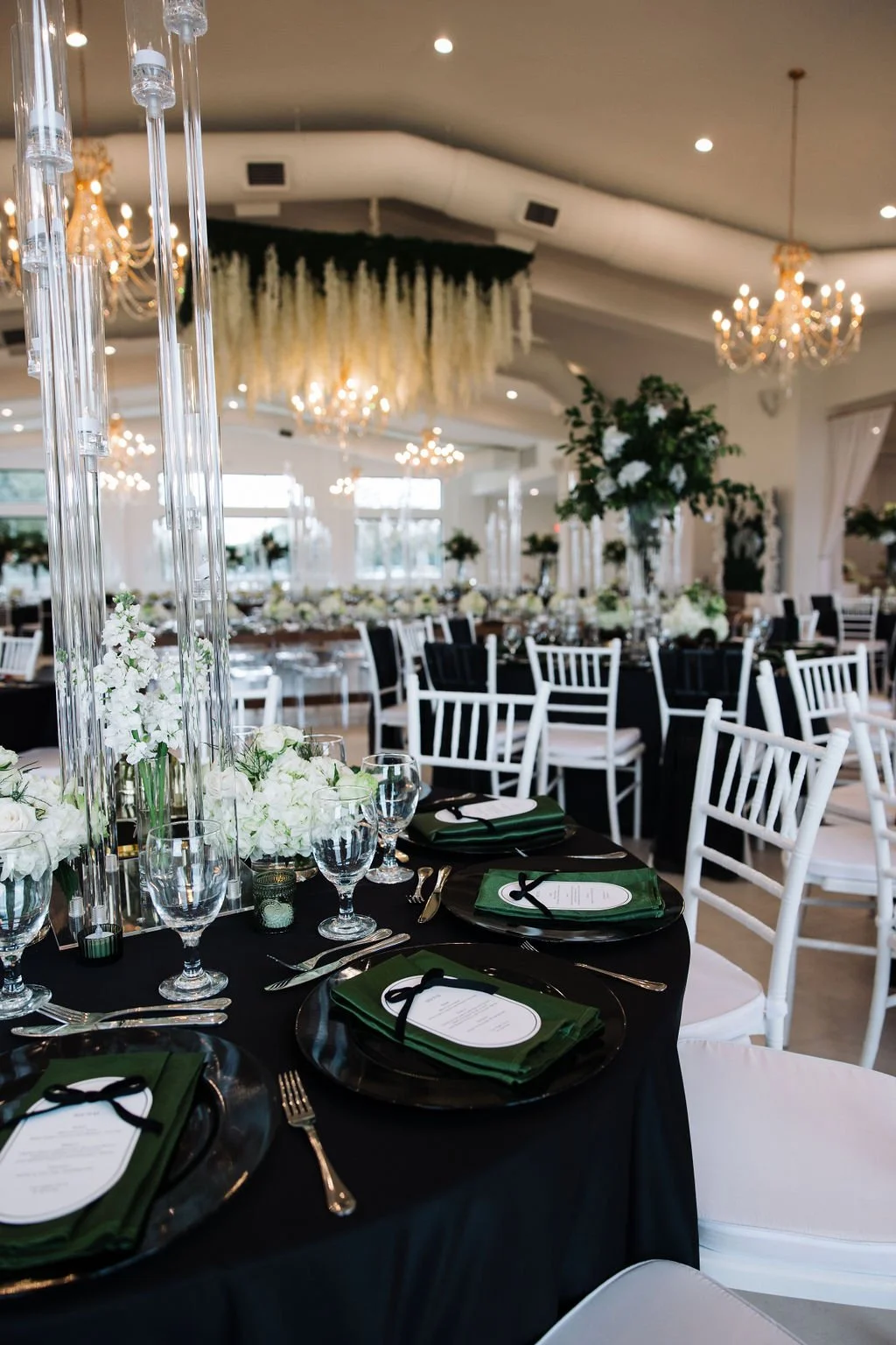Elegant indoor reception hall decorated with white and black color scheme, floral centerpieces, tall glass candleholders, and chandeliers.
