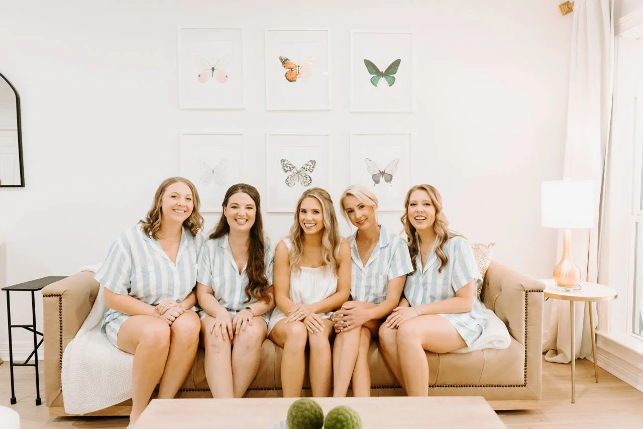 Four bridesmaids and a bride sitting on a beige couch in a bright, modern living room with butterfly artwork on the wall behind them.
