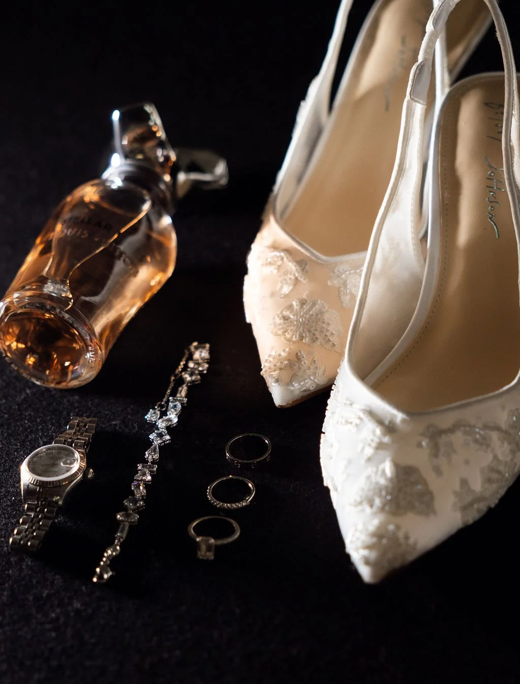 Wedding accessories including white embroidered shoes, a perfume bottle, a watch, rings, a bracelet, and a bracelet with gemstones, all laid out on a dark surface.