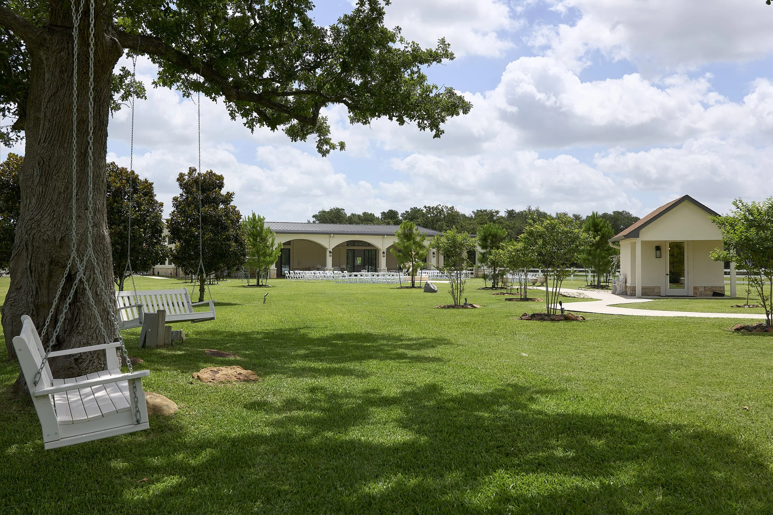 A peaceful outdoor garden area with white benches, a large tree with a swing, and a building with patio seating under a partly cloudy sky.