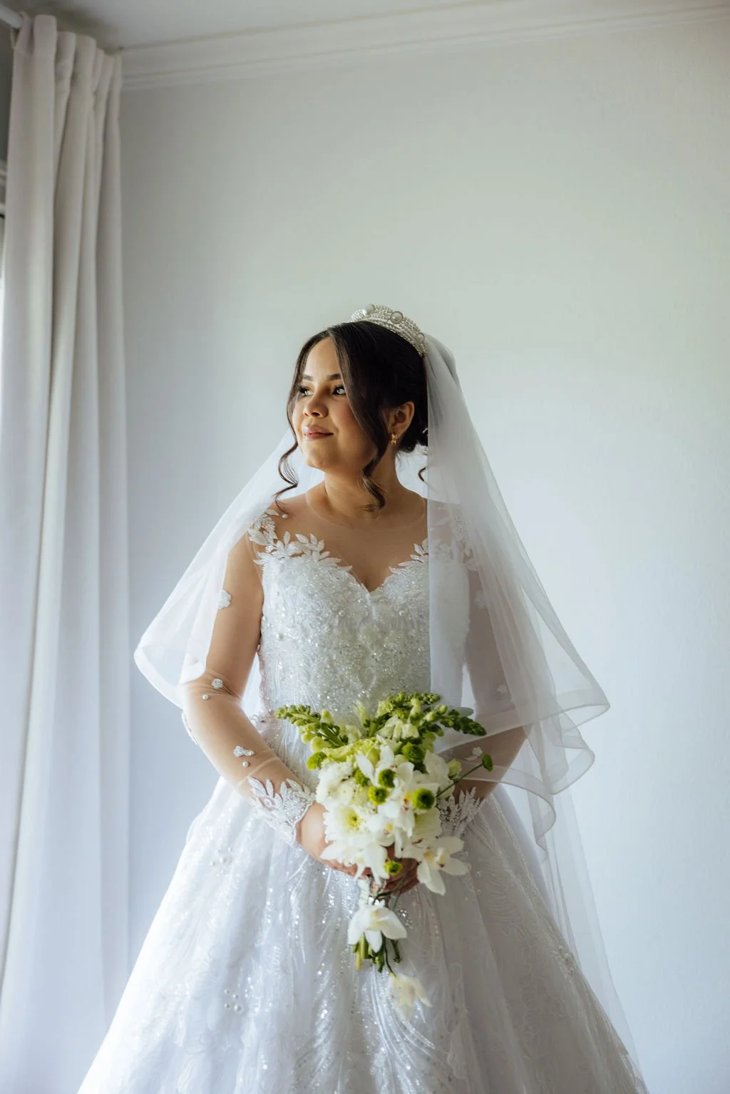 A bride in a white wedding gown with lace and bead details, holding a bouquet of white and green flowers, looking to the side with a gentle smile, standing indoors in the bridal suite near a window with curtains.
