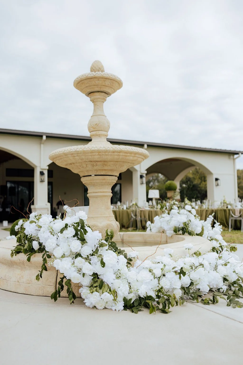 A cream-colored stone fountain decorated with white flowers and greenery, set outdoors with archways and tables in the background at an outdoor wedding reception.