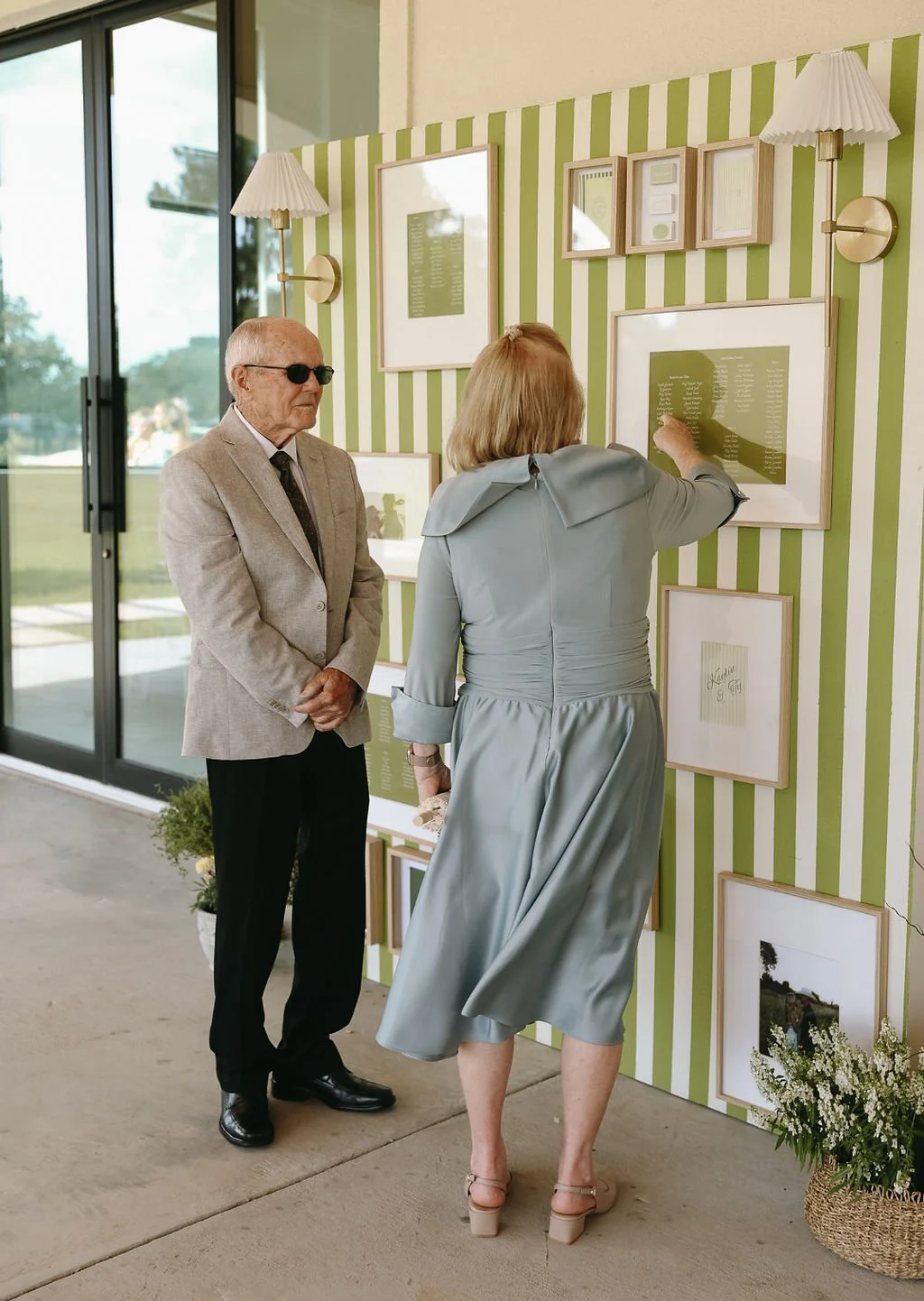 An elderly man with sunglasses and a woman looking at a framed display of a seating chart on a green and white striped wall while enjoying the outdoors at a wedding cocktail hour.