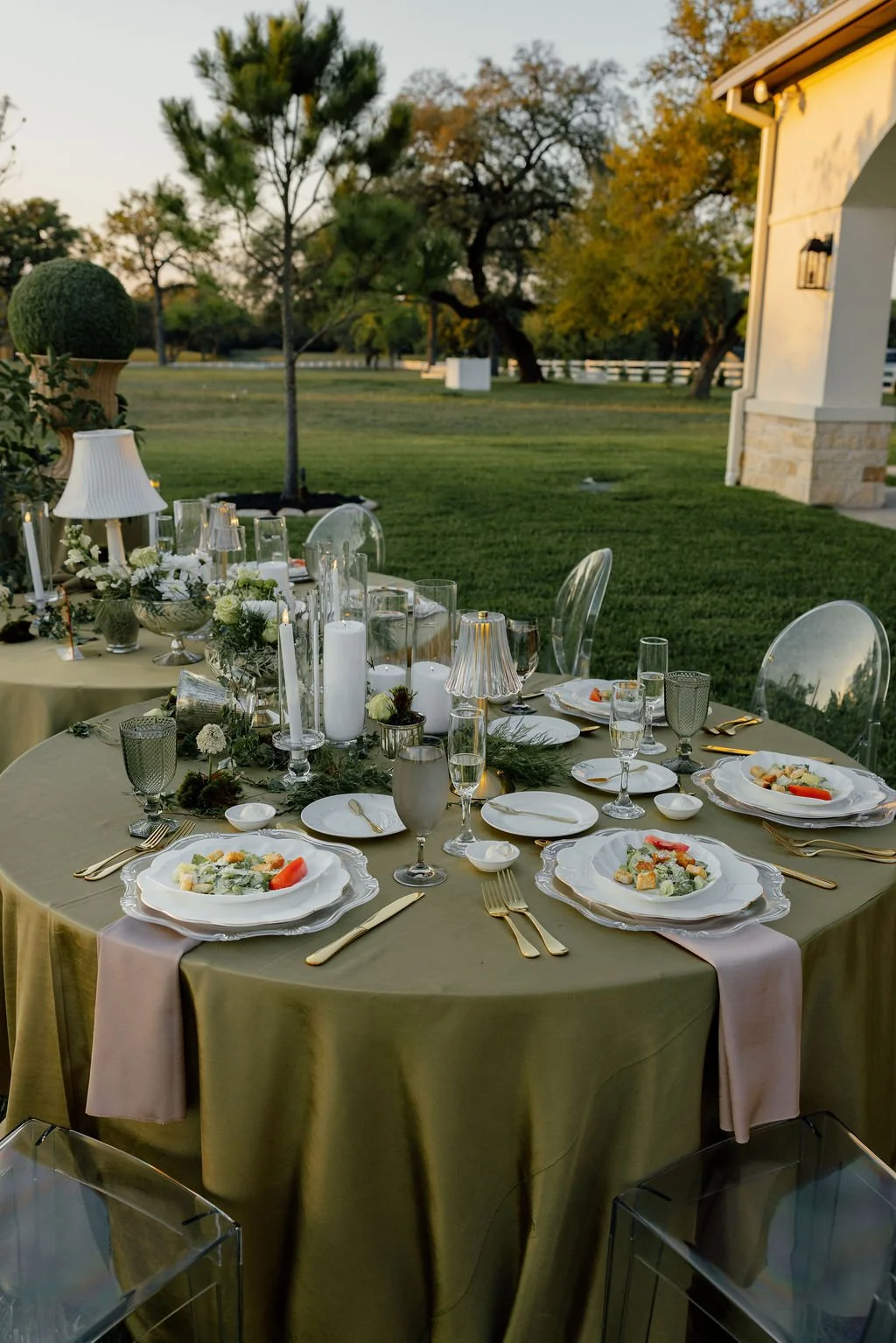 Outdoor dinner setup with a round table covered in a gold tablecloth, set with white plates, gold utensils, and glassware, surrounded by transparent chairs, with candles and floral centerpieces, in a grassy yard with trees and a house in the backgrou