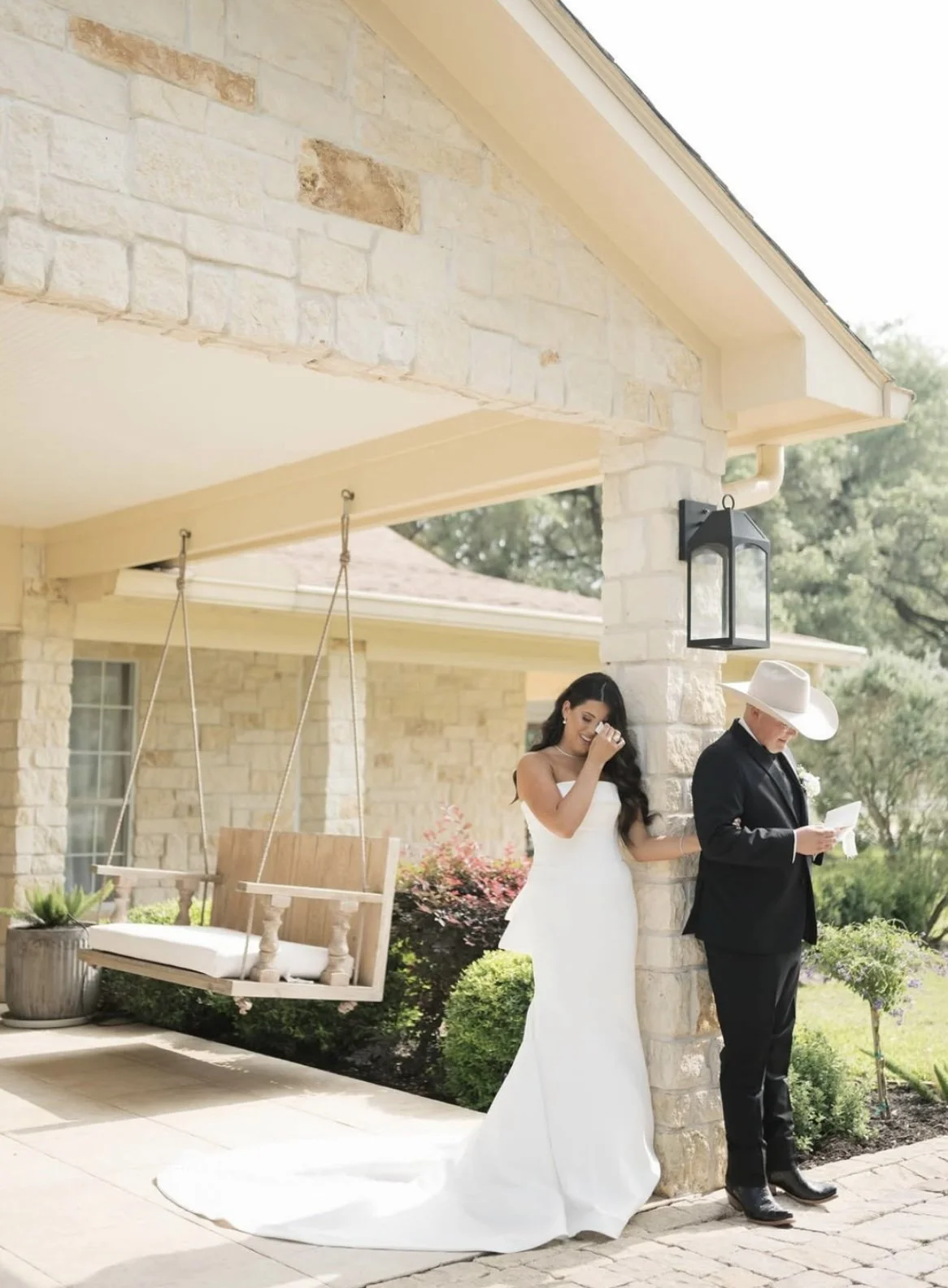 A bride in a white wedding gown and a man in a black suit and white hat are standing outside next to a stone pillar, with the bride laughing and the man reading a card. There is a porch swing hanging nearby and plants surrounding the area.