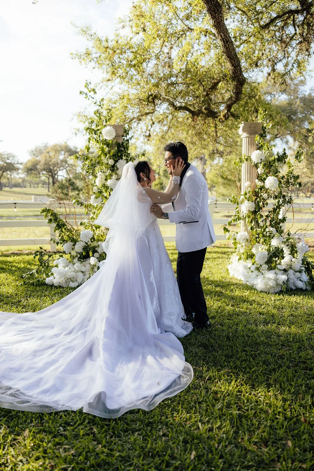 A bride and groom at their outdoor wedding ceremony, standing under a floral arch with white flowers and greenery, embraced and smiling at each other as sunlight filters through trees.