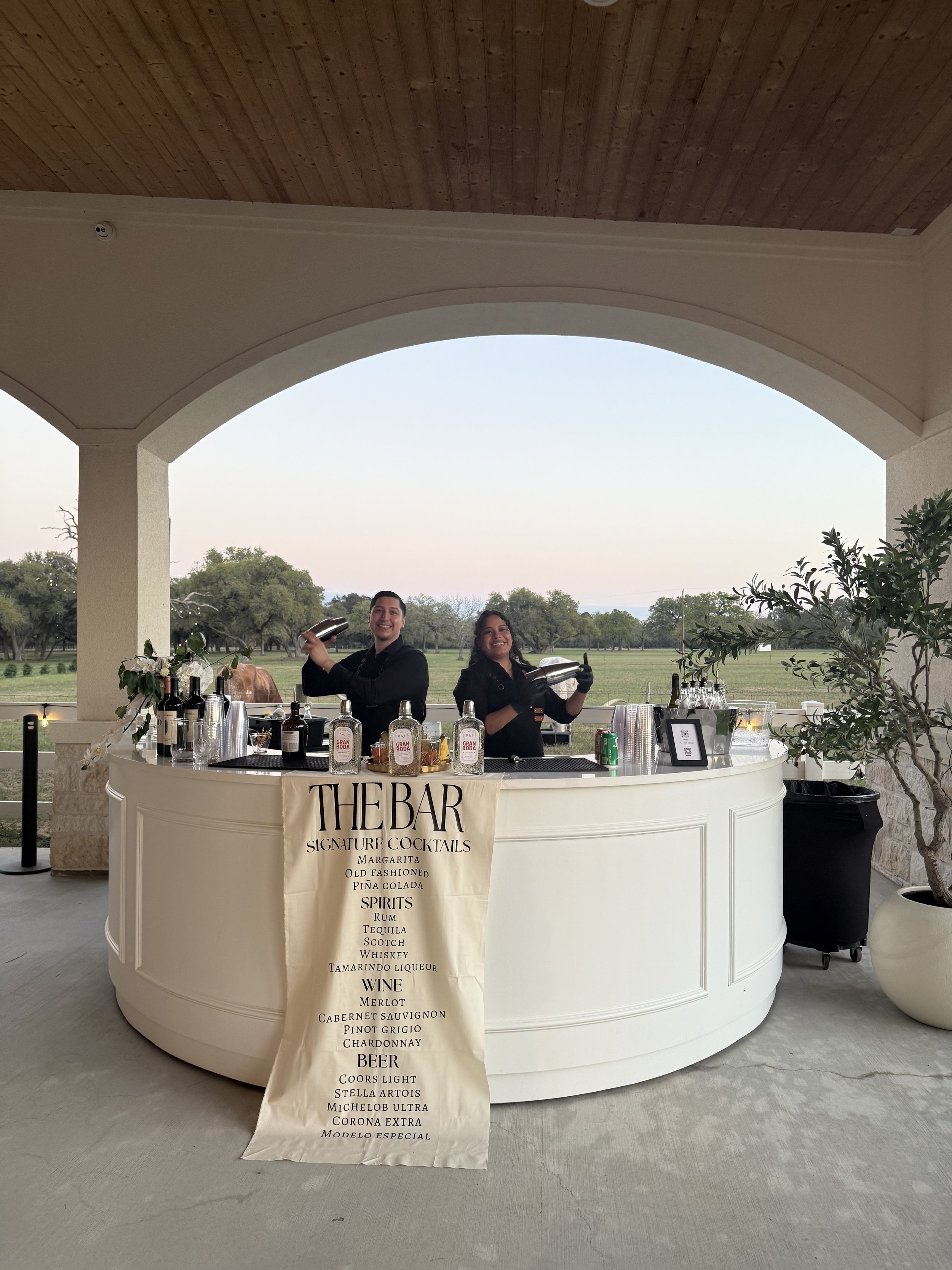 A white bar with bottles of alcohol, mixers, and bar tools. Two smiling bartenders in black attire are behind the bar, holding cocktail shakers. A large sign in front lists drink options including margarita, old fashioned, piña colada, spirits, wine,