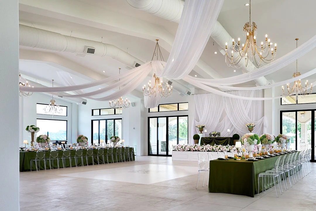 Elegant wedding reception hall with high ceilings, chandeliers, draped white fabric, and floral centerpieces on long tables covered in green tablecloths.