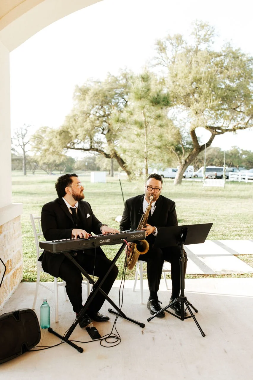 Two men dressed in black suits jazz musicians playing keyboard and saxophone on a porch outdoors, with trees and a grassy field in the background.