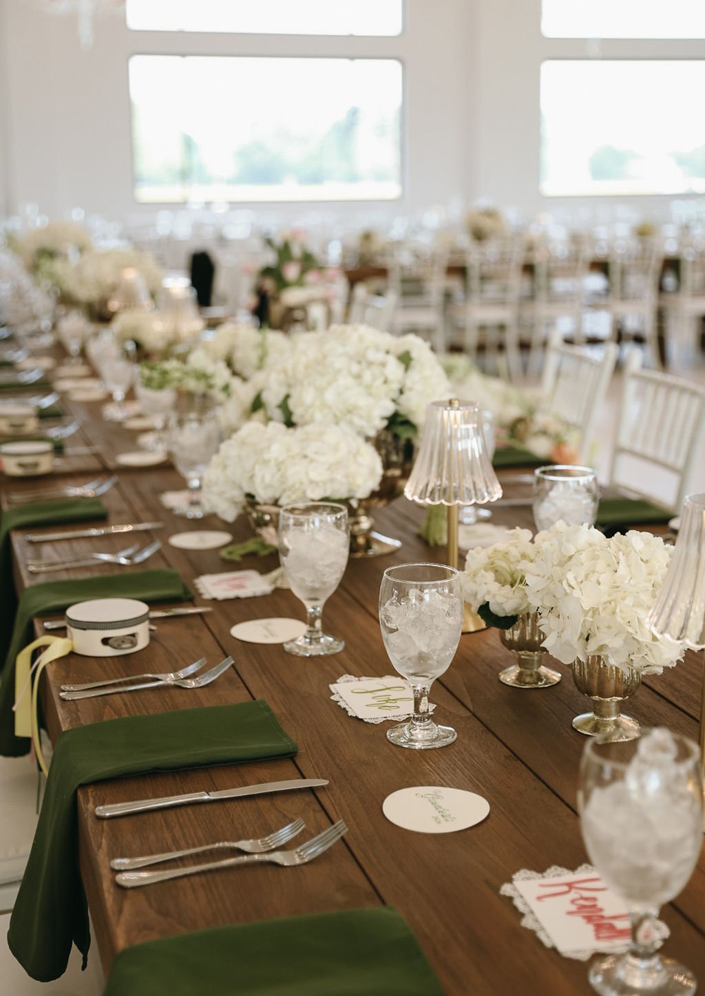 A decorated table set for a wedding at Ro Manor with white floral centerpieces, glassware, silverware, green napkins, and small lamps.