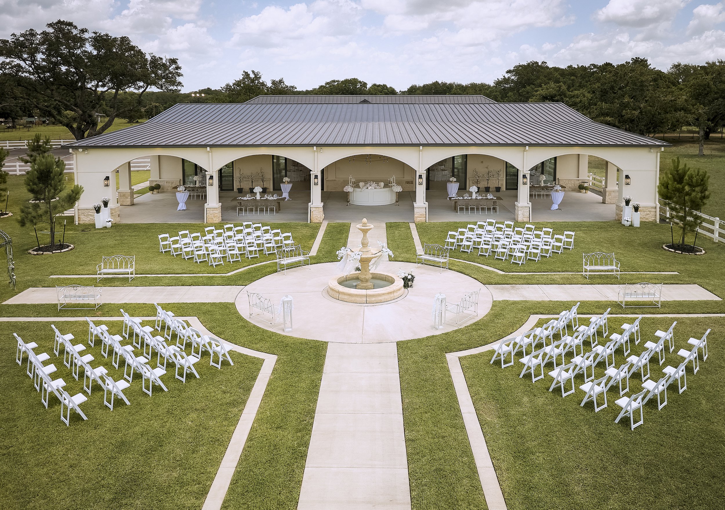 An outdoor wedding setup with white chairs arranged in rows facing a small fountain and a covered pavilion decorated with white flowers and table arrangements. There are trees and a cloudy sky in the background.