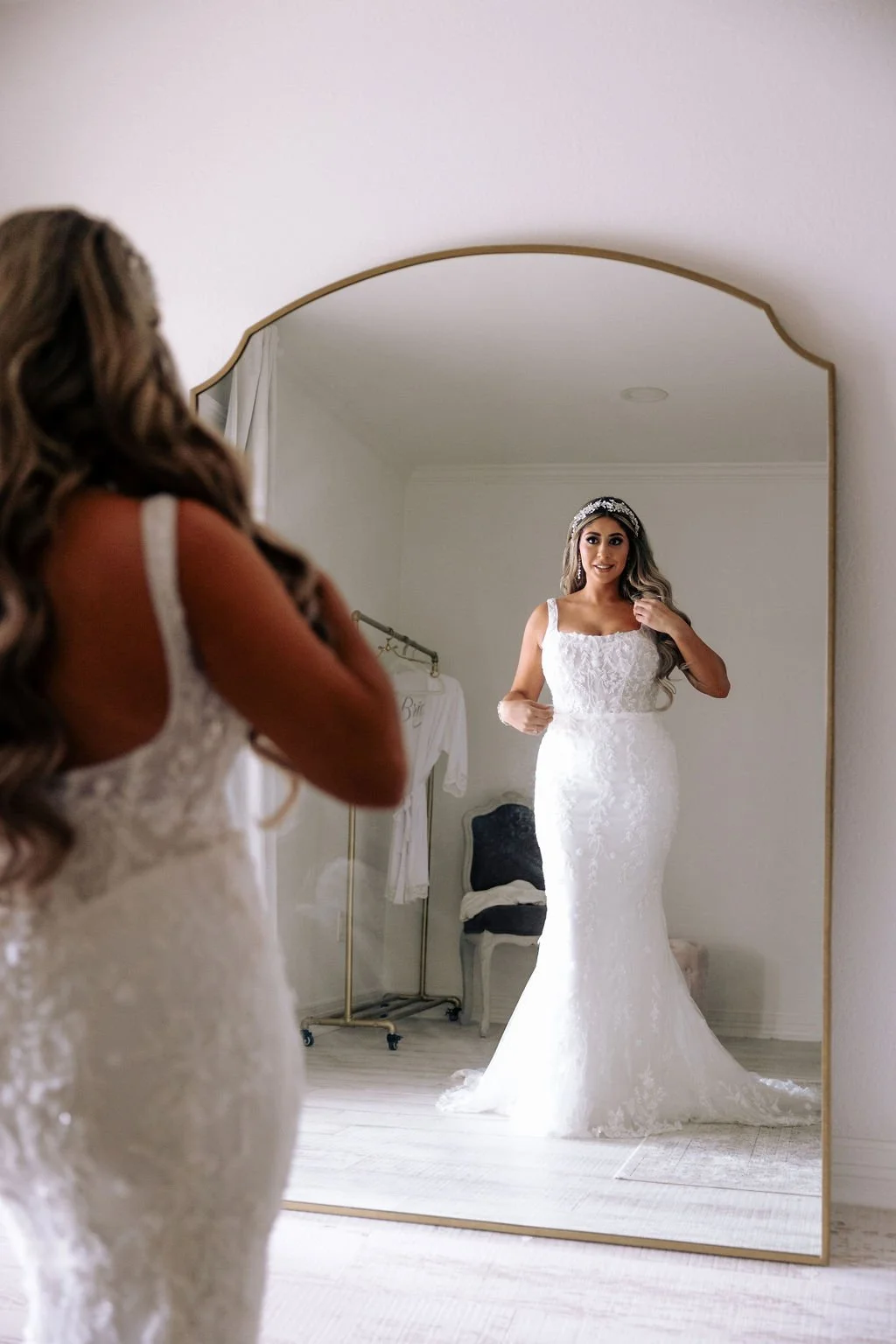 Brunette bride in a white lace wedding dress with a beaded headband looking at herself in a large mirror.