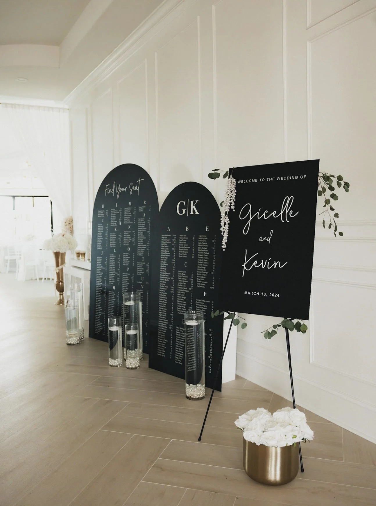 Wedding welcome sign and seating chart displays with black boards, greenery, and white flowers in vases inside a bright, elegant venue.