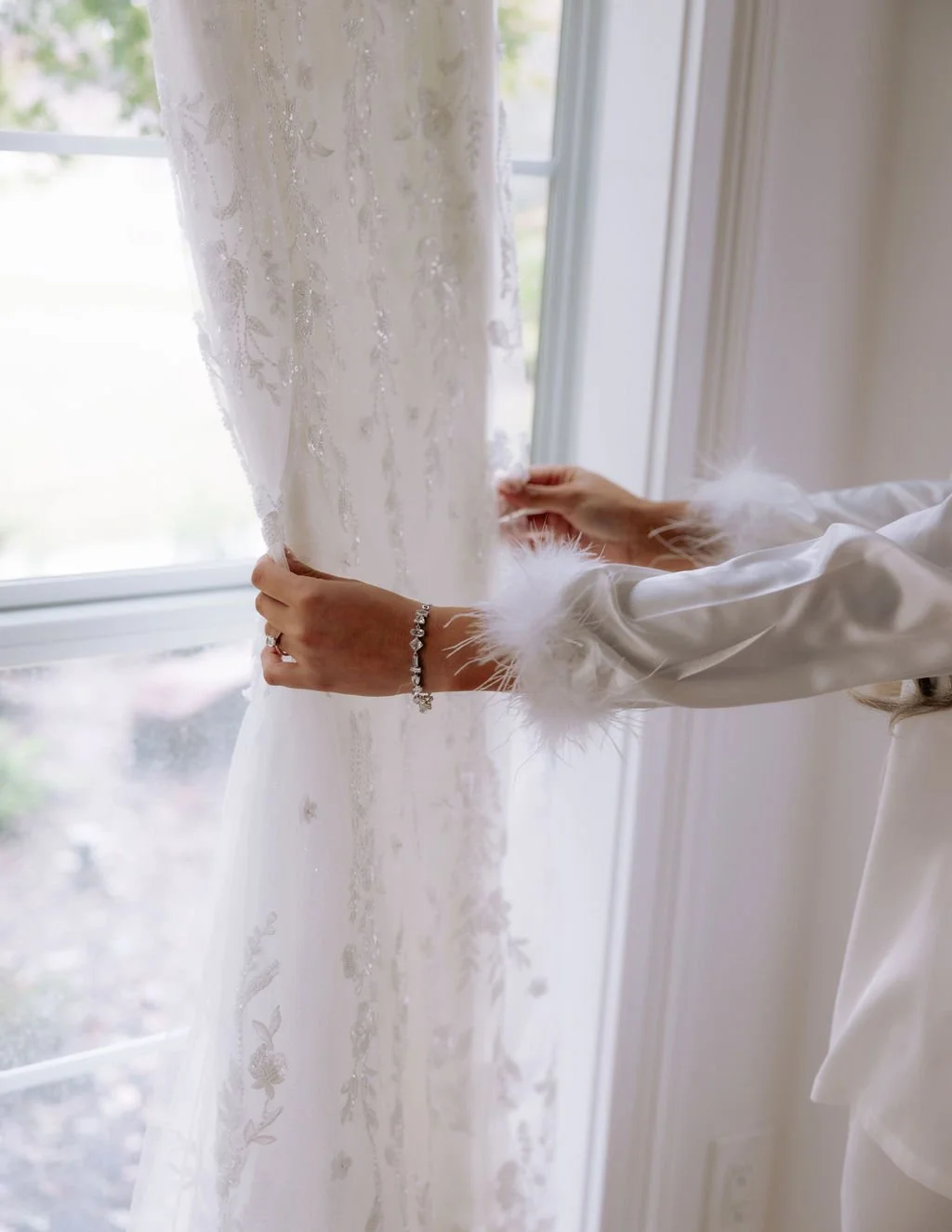 Bride in a white satin robe with feather trim adjusting a lace wedding dress in front of a large bay window.