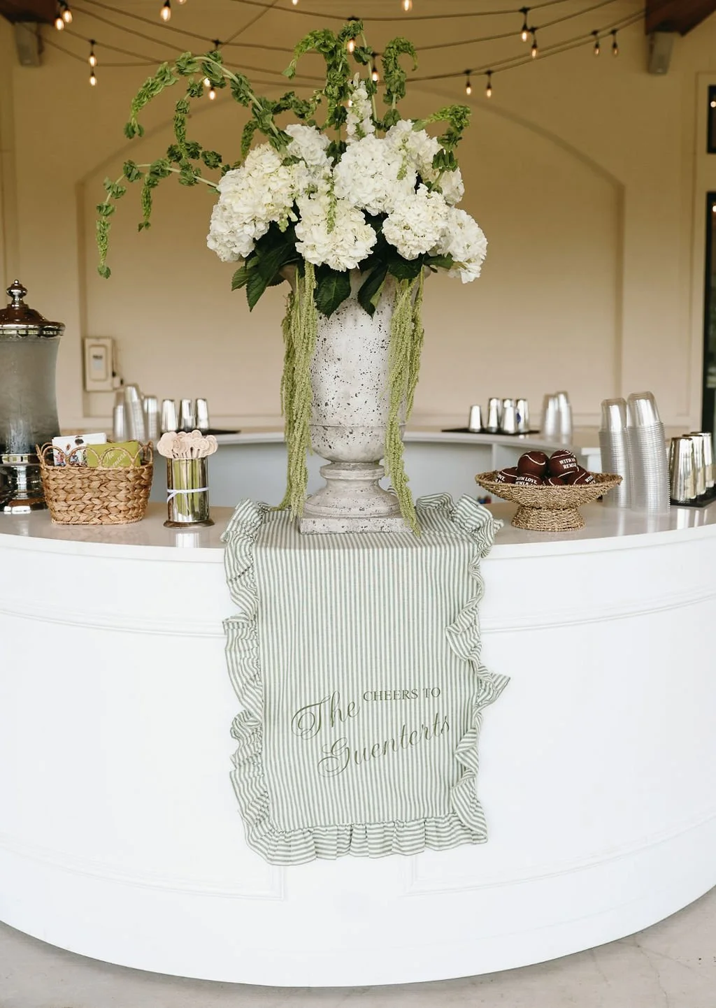 Large white floral arrangement with hydrangeas and greenery in an ornate white vase placed on a striped fabric cloth on a white curved counter. Behind the counter, there are disposable cups and small containers, indicating a celebratory setting.