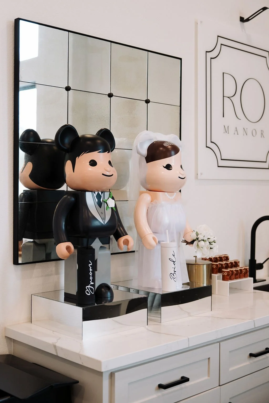 Decorative figurines of a bride and groom on a countertop, with a mirror reflecting the groom figure, flower decor, and a sign that says "RO MANOR."