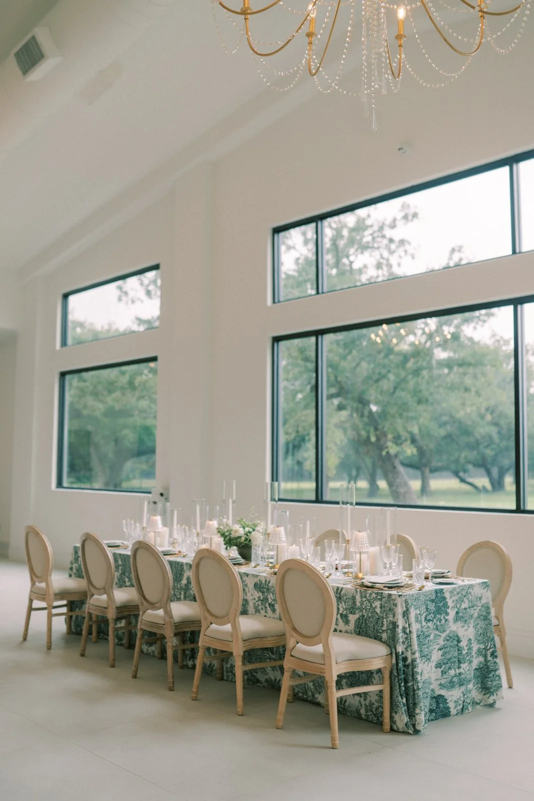 Elegant dining room table set for a wedding, with a patterned tablecloth, chairs, and candle-lit centerpieces, large windows revealing greenery outside, and a large gold chandelier hanging from the ceiling.