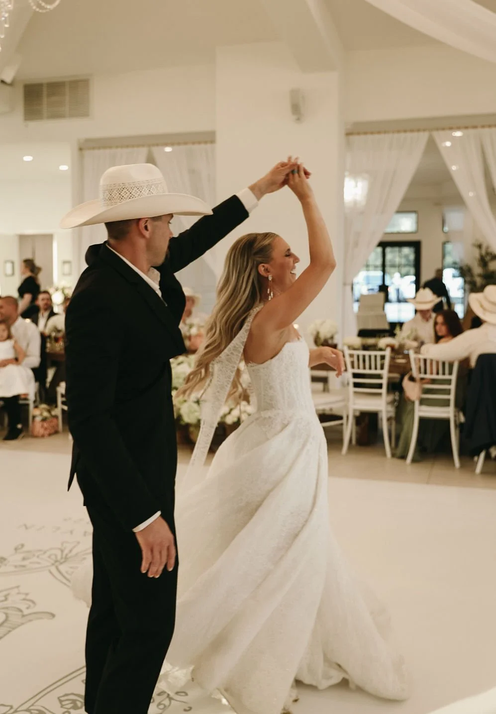 Couple dancing at wedding reception, the bride in a white wedding dress and the groom in a black suit with a large white hat, surrounded by guests seated at tables in a decorated venue.