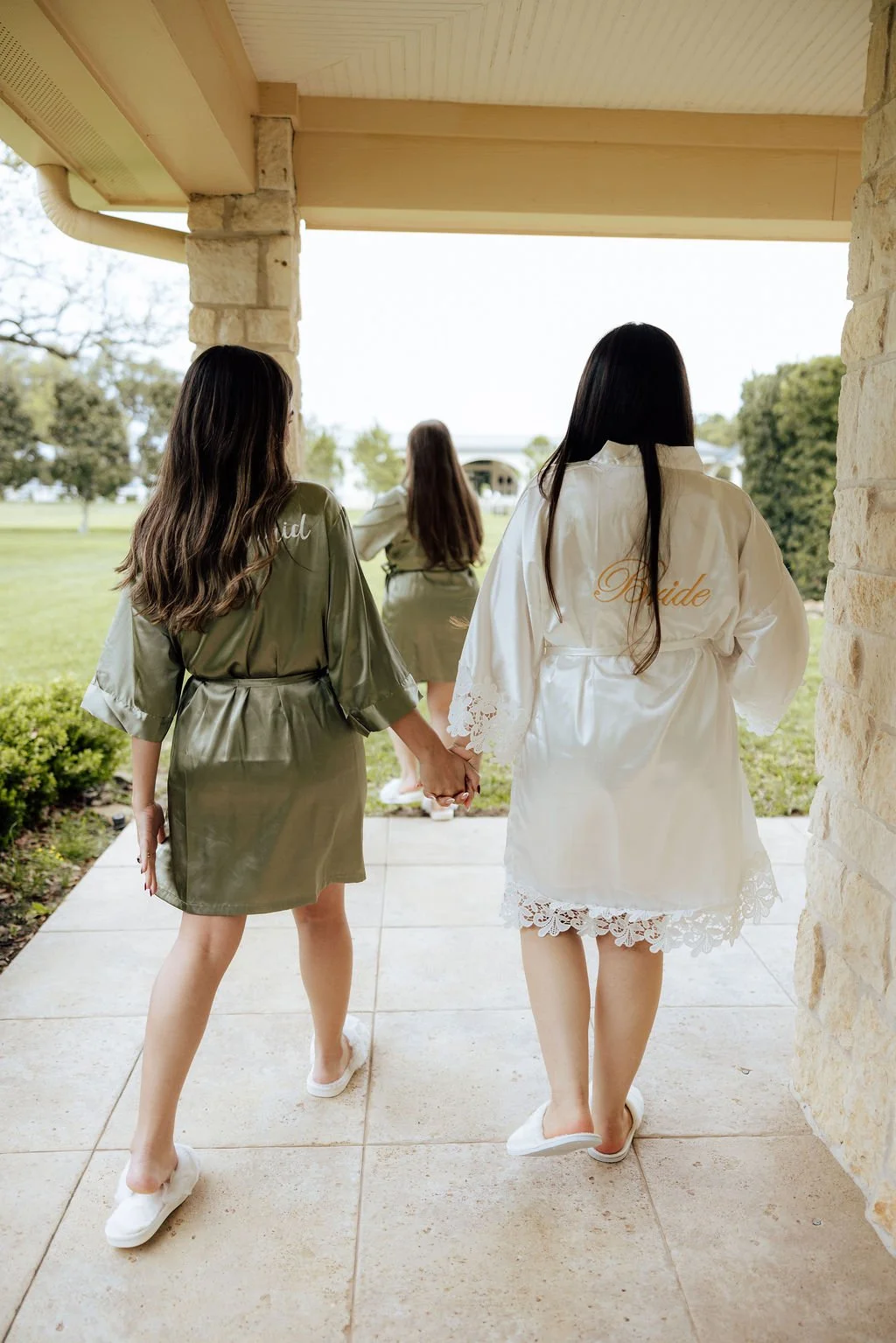Wedding morning. Three women in bridal satin robes walk hand in hand on a porch, with two women wearing white robes labeled 'Bride' and one in an olive-green robe, holding hands as they approach outdoor surroundings. 