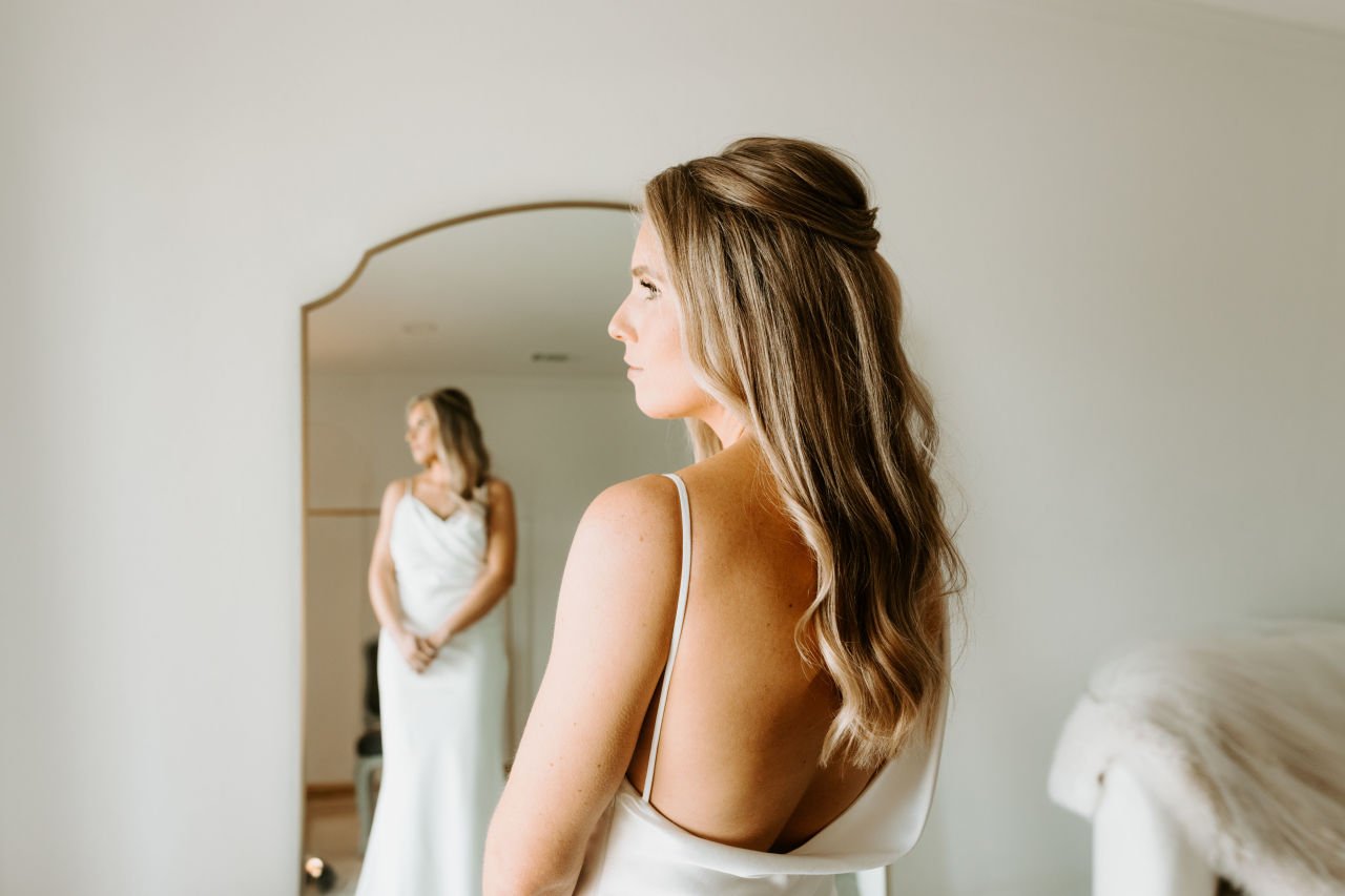 A bride with wavy brown hair looking at her reflection in a mirror at Ro Manor's bridal suite, wearing a white silk dress.