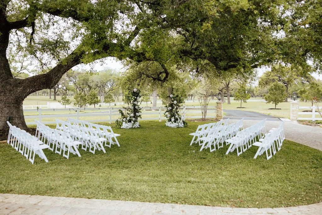 Outdoor wedding ceremony setup with white garden chairs arranged on green grass under a large tree, decorated archway in the background, on a sunny day.