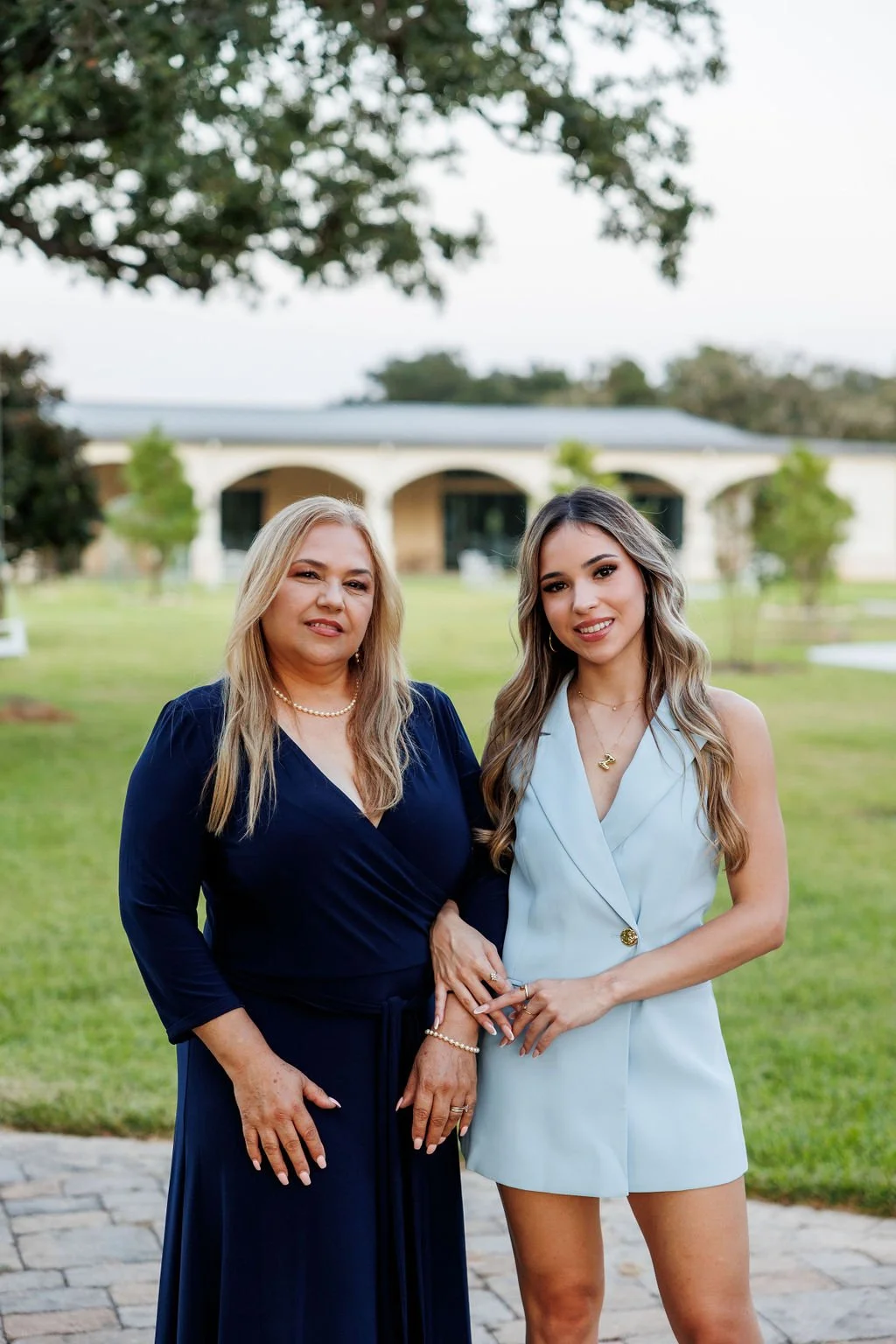 Two women standing outside on grass, one older with long gray hair wearing a navy blue dress, and one younger with long wavy blonde hair wearing a light blue sleeveless blazer dress, both smiling and holding hands. In the background, there is a large white building with arches and trees.